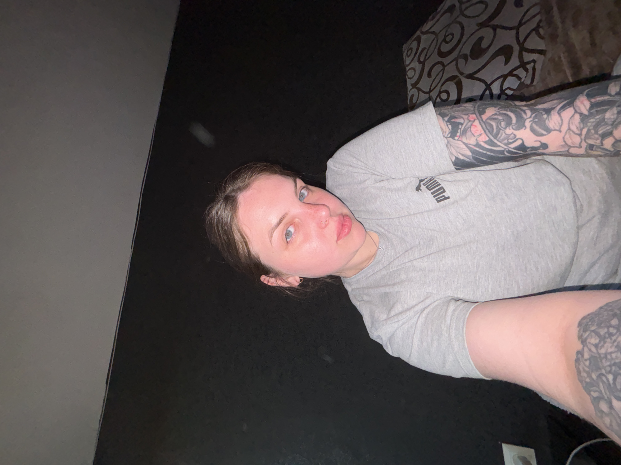 A self-portrait captured from a low angle shows a young woman with light skin, blue eyes, and brown hair, looking directly at the camera with a neutral expression. She has a subtle nose piercing. She is wearing a light gray, short-sleeved t-shirt, with the brand name "PUMA" visible on the fabric of her left sleeve. Both her forearms are extensively covered with intricate tattoos featuring black and red patterns, including floral and traditional designs.
The background is dim, suggesting an indoor setting in low light or at night. On the viewer's left, a light gray wall or vertical surface is partially visible. The rest of the background is a dark, undefined area, with a portion of brown, patterned upholstery visible behind her upper right arm. The image is taken in Chornomorskoe, Ukraine.