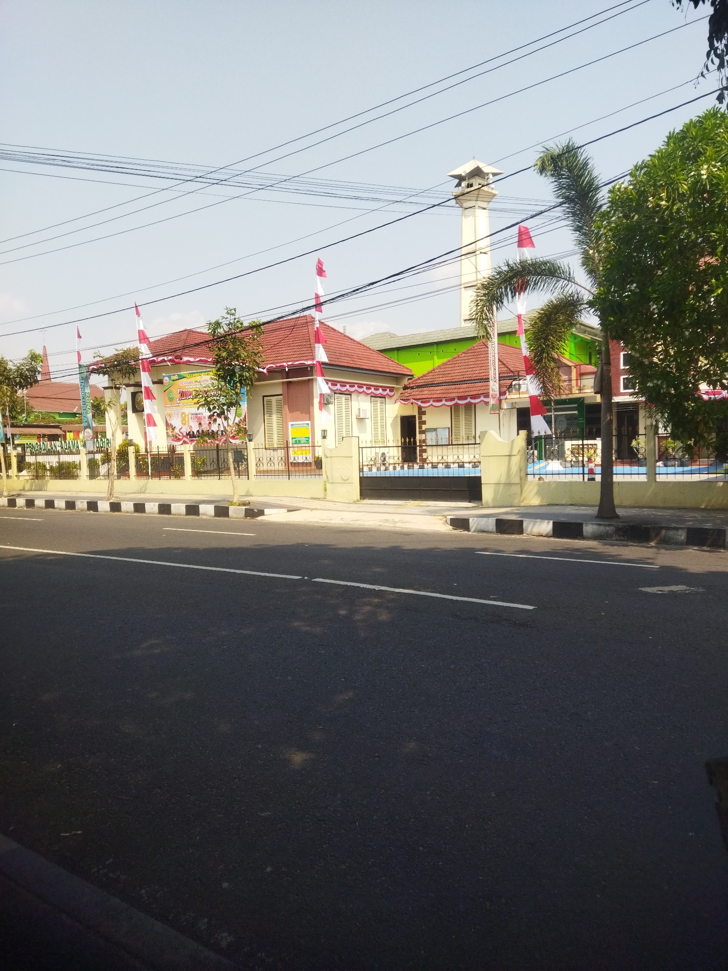 The image shows the front of the Religious Affairs Office (Kantor Urusan Agama) in the city of Wonogiri, Indonesia. The building has a red tiled roof, with white and red Indonesian flags displayed along the facade. A large banner with text and images is also visible on the building. In front of the building is a paved sidewalk with black and white striped borders. A road with white lane markings runs in front of the sidewalk. A tall, square minaret with a decorative top stands behind the building, to the right of the frame. Green trees are visible on the right side of the image. The sky is clear and blue. The overall scene depicts an official building in a city setting under a clear sky.