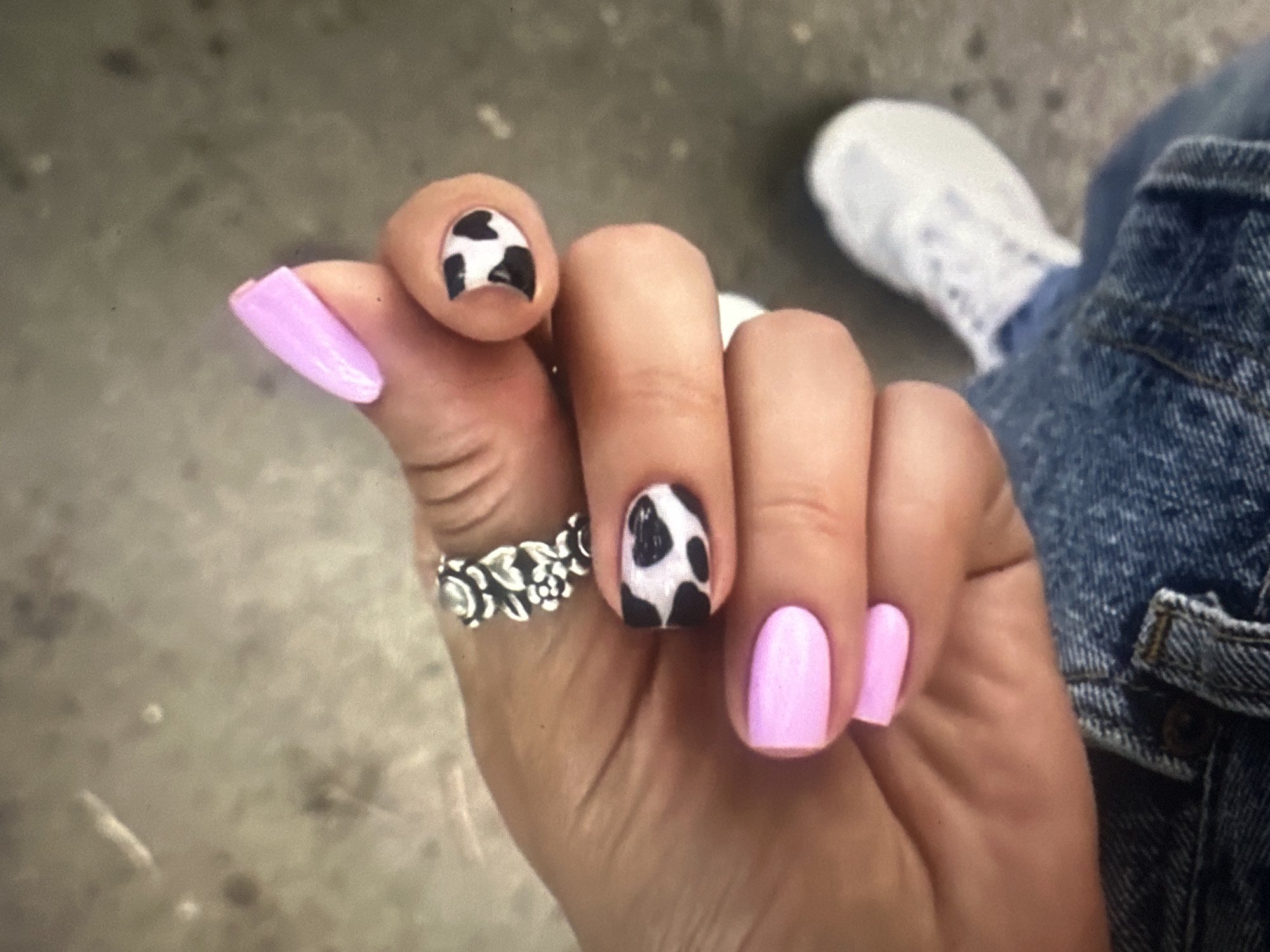 A close-up shot focuses on a person's hand with a unique manicure. The nails are adorned with a pink base coat and feature a cow print design on some of the fingernails, specifically the index finger and thumb, which showcase black irregular spots against a white background. A silver ring with a floral design is worn on the ring finger. The person is wearing blue denim jeans and a white sneaker is visible in the background. The setting appears to be an outdoor urban environment, possibly a sidewalk or pavement.