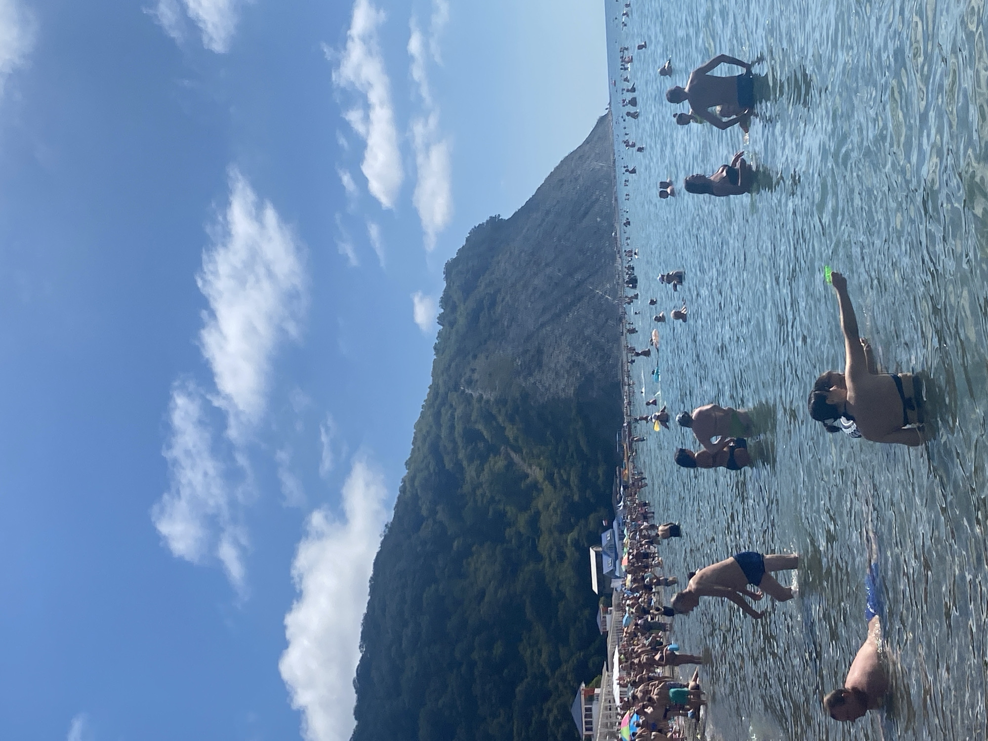 This image depicts a crowded beach in Arkhipo-Osipovka, Russia, under a bright blue sky with scattered white clouds. A steep, tree-covered mountainside dominates the background, sloping down to the sea. The water is filled with numerous people swimming, wading, and interacting. In the foreground, a man is lying in the shallow water, and another man is wading through the water. Further out, several groups of people are visible, with some appearing to be playing or socializing. Along the shoreline, tents, umbrellas, and buildings are present, indicating a developed tourist area. The overall scene conveys a bustling summer day at a popular seaside resort.