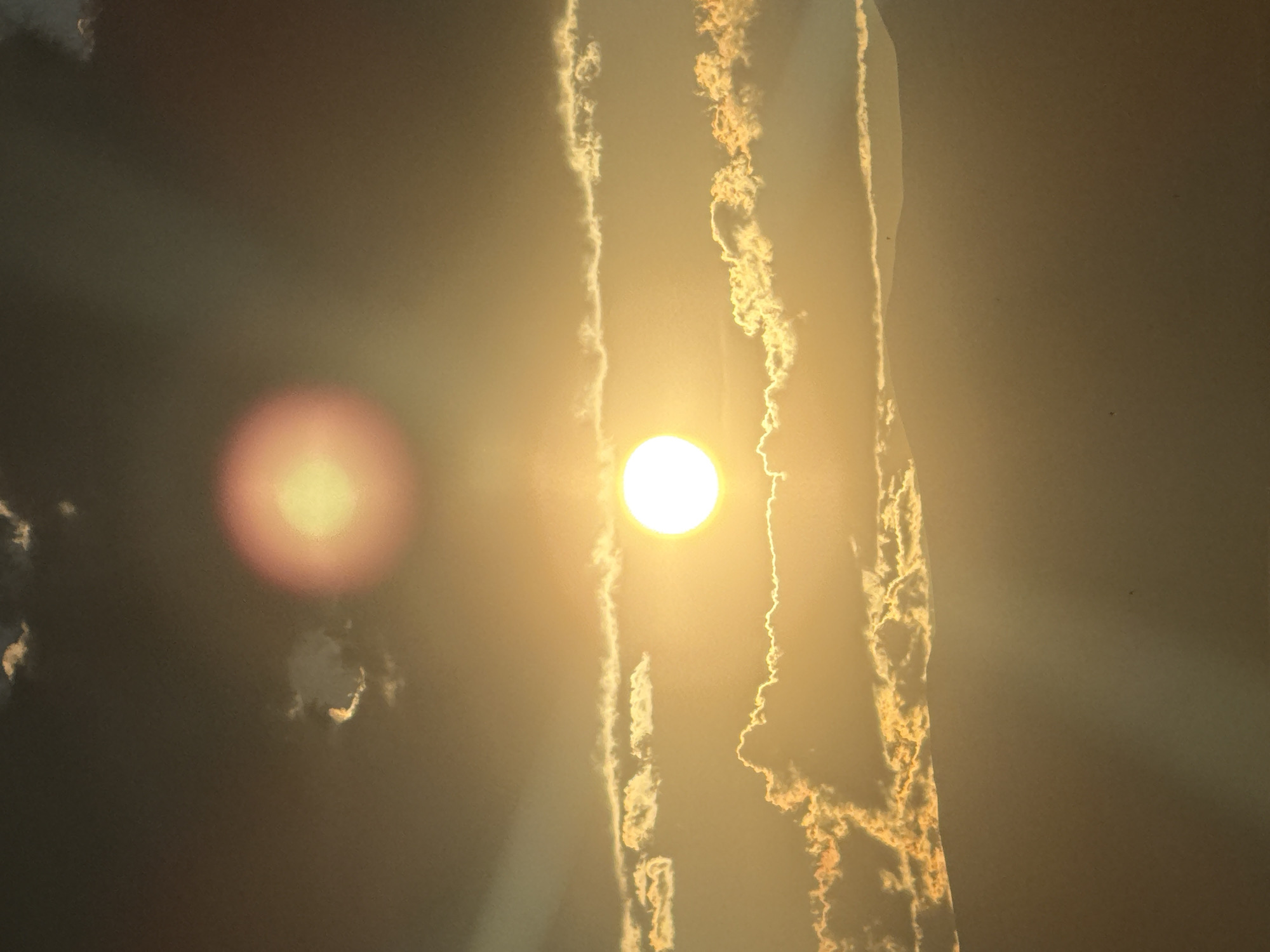 The image captures a vibrant and hazy sky, likely at sunrise or sunset, over Abovyan, Armenia. The central subject is the bright, circular sun, positioned slightly to the right of the image center, casting strong, golden rays through the atmosphere. To the left of the sun, a distinct, soft, red-orange circular lens flare is visible, suggesting the angle of the sun relative to the camera lens.
The sky is characterized by a warm, golden-brown hue, indicative of atmospheric haze or dust. Several prominent, elongated vertical formations, resembling wispy clouds or contrails, stretch across the frame. These trails are brightly illuminated by the sun, appearing white and golden with intricate, feather-like textures, contrasting against the darker, hazy sky.
The scene conveys a sense of serene natural beauty and atmospheric depth. There are no visible people, objects besides the sun and atmospheric elements, or any discernible text. The image focuses entirely on the celestial display and the warm, diffused light.