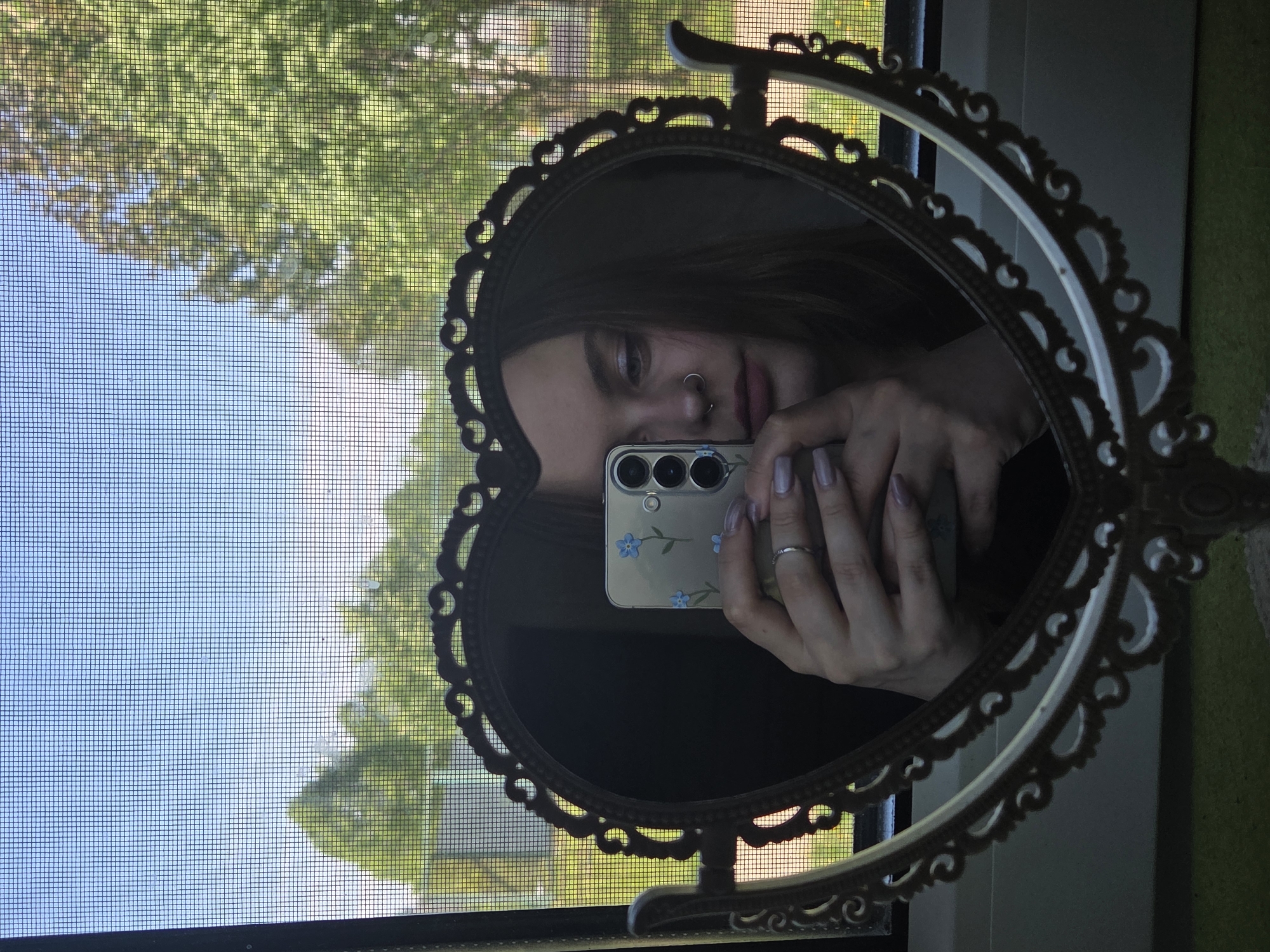 A person is reflected in a heart-shaped ornate mirror, holding a smartphone up to their face to take a picture. The phone has a clear case with blue floral designs and multiple camera lenses visible. The person has fair skin, a ring on their finger, and visible nose piercings. The mirror is positioned in front of a window with a screen, which shows an exterior view of trees and sky. A portion of a green wall or surface is visible on the right side of the mirror. The location is likely indoors, positioned near a window.
