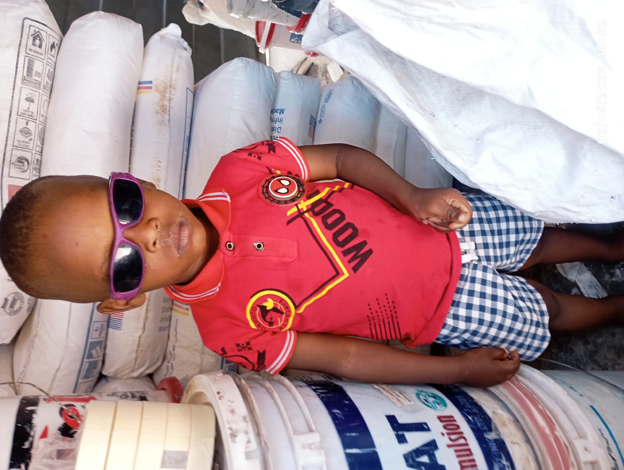 A young boy with dark skin and short hair lies down on a stack of white bags and paint buckets. He wears purple sunglasses, a red t-shirt with "WOOD" printed on it, and blue and white checkered shorts. The bags appear to be sacks of ingredients, possibly for building material. The paint buckets contain the word "emulsion". The date "02/05/2026 10:02" is digitally displayed in the upper right of the frame. The image was taken in the city of Jos, Nigeria.