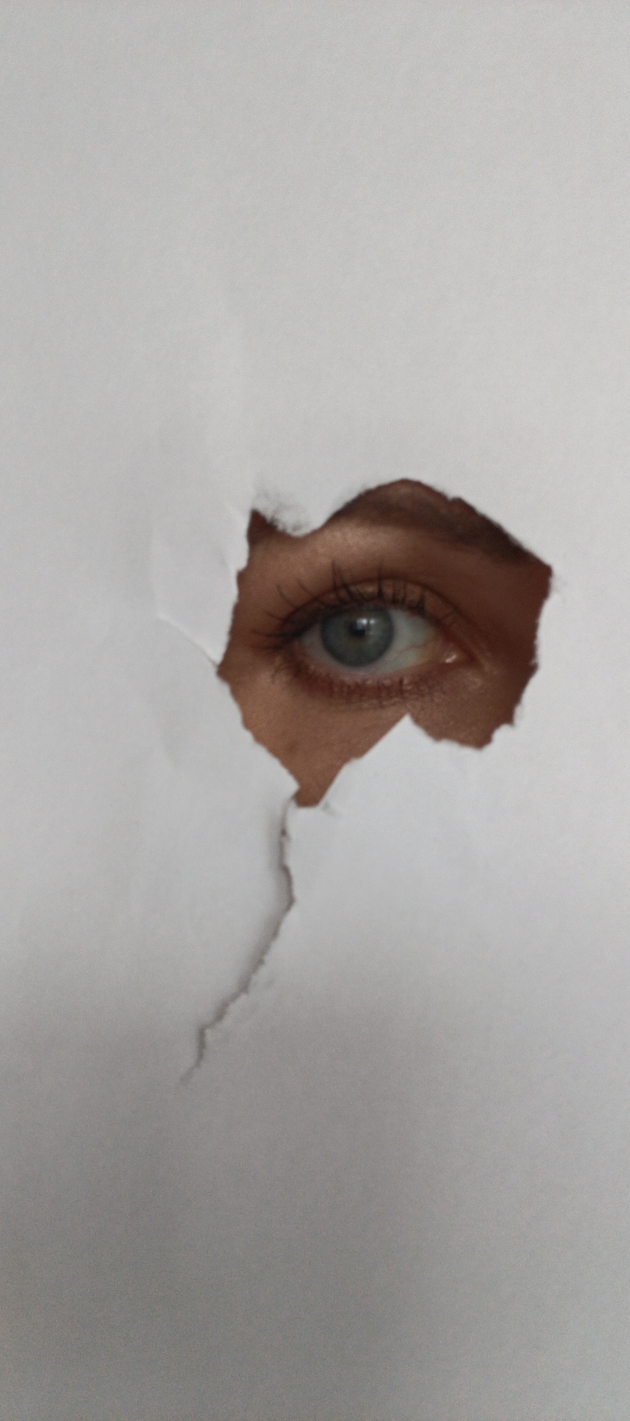 A close-up, vertical photograph captures a human eye peering through an irregularly torn hole in a sheet of plain, light-colored paper. The eye, appearing to be the right eye of a person, features a distinct light blue-grey iris with a visible, round pupil and a small catchlight. Long, dark eyelashes are prominent around the eye. The skin visible around the eye and eyebrow through the rough opening is a warm, light-brown tone.
The white or very light grey paper dominates the foreground, with its jagged, torn edges forming the aperture through which the eye is seen. The texture of the paper is subtly visible. The direct gaze of the eye suggests an act of observation or peeking. No specific time of day or weather conditions are discernible from the image. There is no visible text. The location context for this media file is Chaikovsky, Russia.