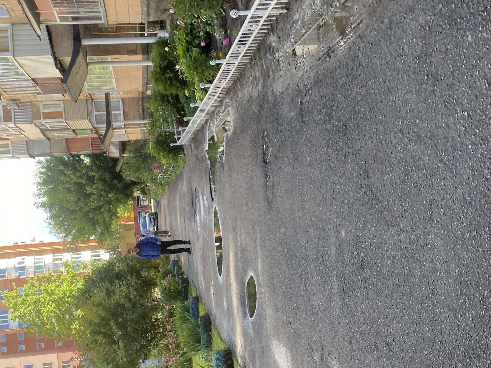The image shows a street scene in Poltava, Ukraine, with a gray asphalt road prominently displayed. Along the side of the road is a combination of green vegetation, including trees and flowers, and a white metal fence. There are puddles on the road. Two people are walking away from the viewer down the road. In the background, residential buildings of varying heights and architectural styles are visible.