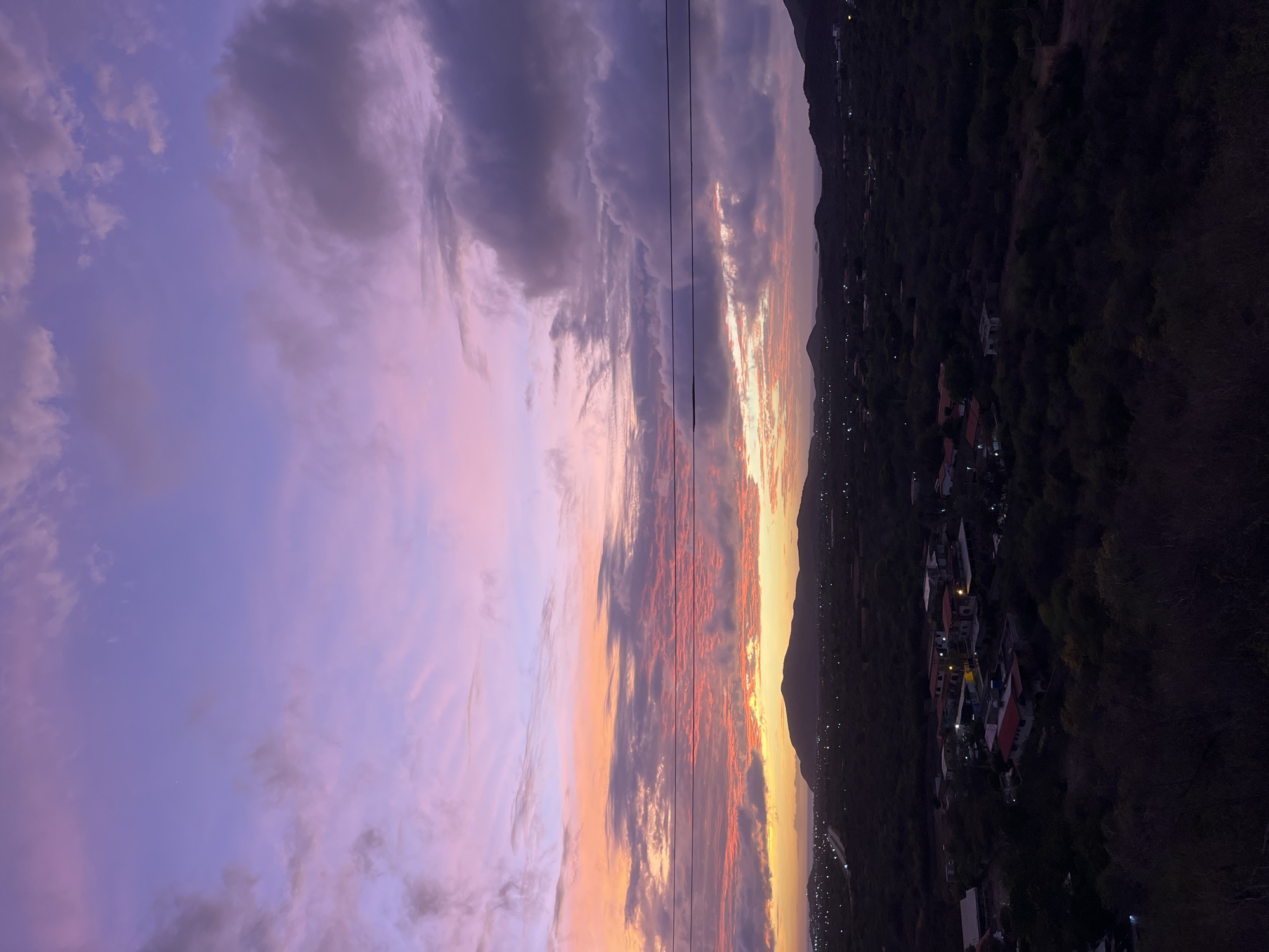 A wide shot captures a twilight scene over the city of Santa Ana, Venezuela. The sky is a gradient of soft purples, pinks, and oranges, with streaks of white clouds. In the distance, a range of dark hills forms the horizon, silhouetted against the vibrant sky. Below the hills, the city lights of Santa Ana are visible as scattered points of light amidst the dark landscape. Power lines are stretched across the frame horizontally, cutting across the sky. The foreground is dominated by dark, dense foliage, with some scattered buildings visible within the greenery. The overall impression is one of a tranquil, twilight setting in a semi-rural area.