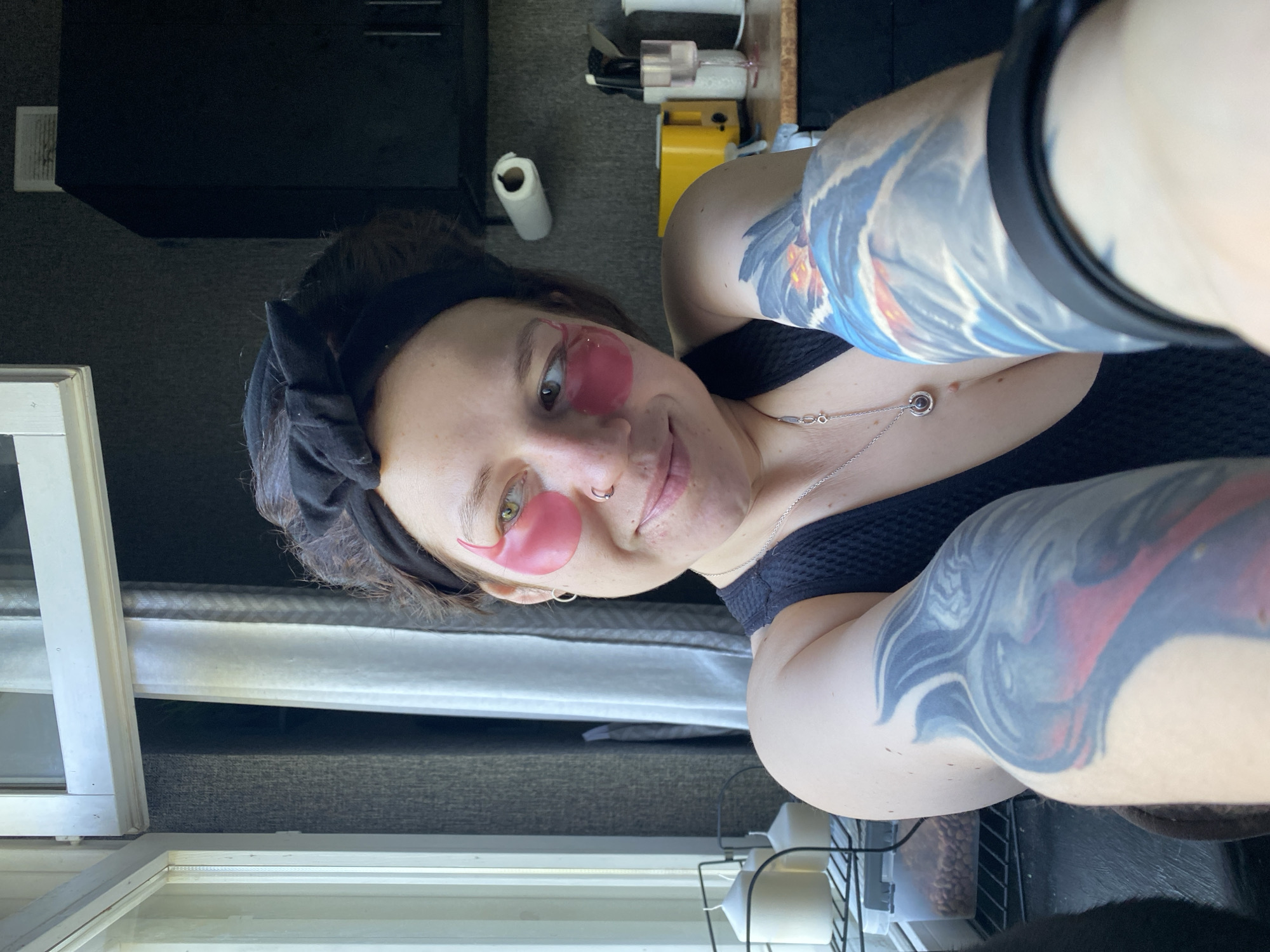 A woman with dark hair pulled back by a black headband and pink under-eye patches is looking to her right, smiling. She is wearing a black tank top and a silver pendant necklace. Her left arm, which is visible in the foreground and features a large blue and red tattoo, is extended towards the camera. She has a nose ring and an earring in her left ear. In the background, a window with white blinds and sheer curtains is to the left, and kitchen cabinetry is visible to the right. A yellow toaster and a roll of paper towels are also on the counter. The image is taken indoors, likely in a kitchen or living area. There is no information available to determine the location in the city of Chaikovsky, Russia.