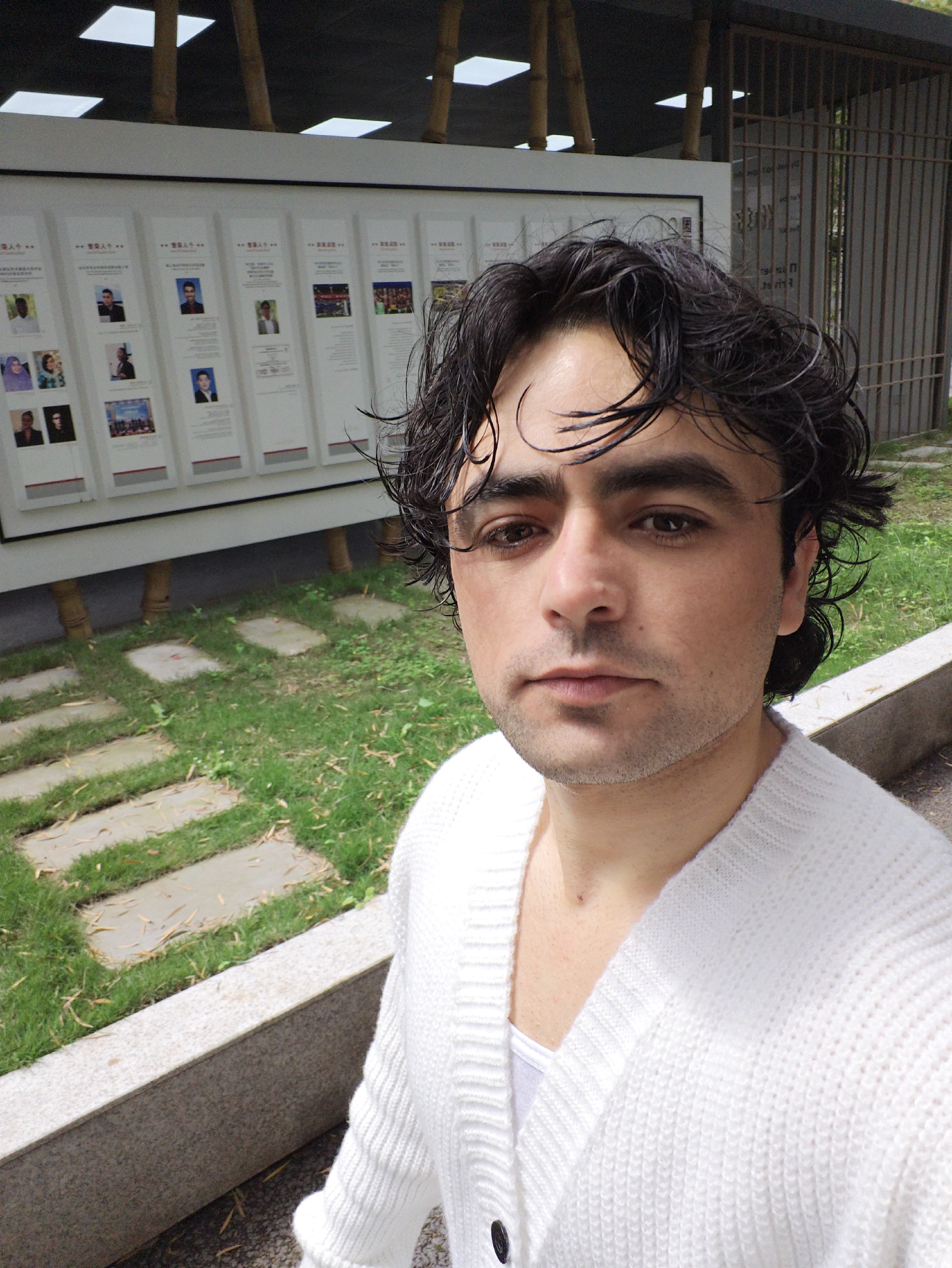 A medium shot captures a man with dark, wavy hair, wearing a white cardigan over a white t-shirt. He is the focal point of the image, positioned in front of a public information board displaying individual photos and brief profiles. Behind the man and the board is a paved area of grass and stone, indicative of a park or public space. A fenced structure and bamboo supports can be seen in the background.
The location is identified as the Chongqing High-tech Zone in China. The scene appears to be outdoors, possibly a memorial or exhibition area within a park, indicated by the public display, manicured grass, and surrounding structures. The man's activity appears to be taking a selfie, as he is directly facing the camera with his body angled slightly. There is no indication of any specific interaction with other people or elements in the environment.