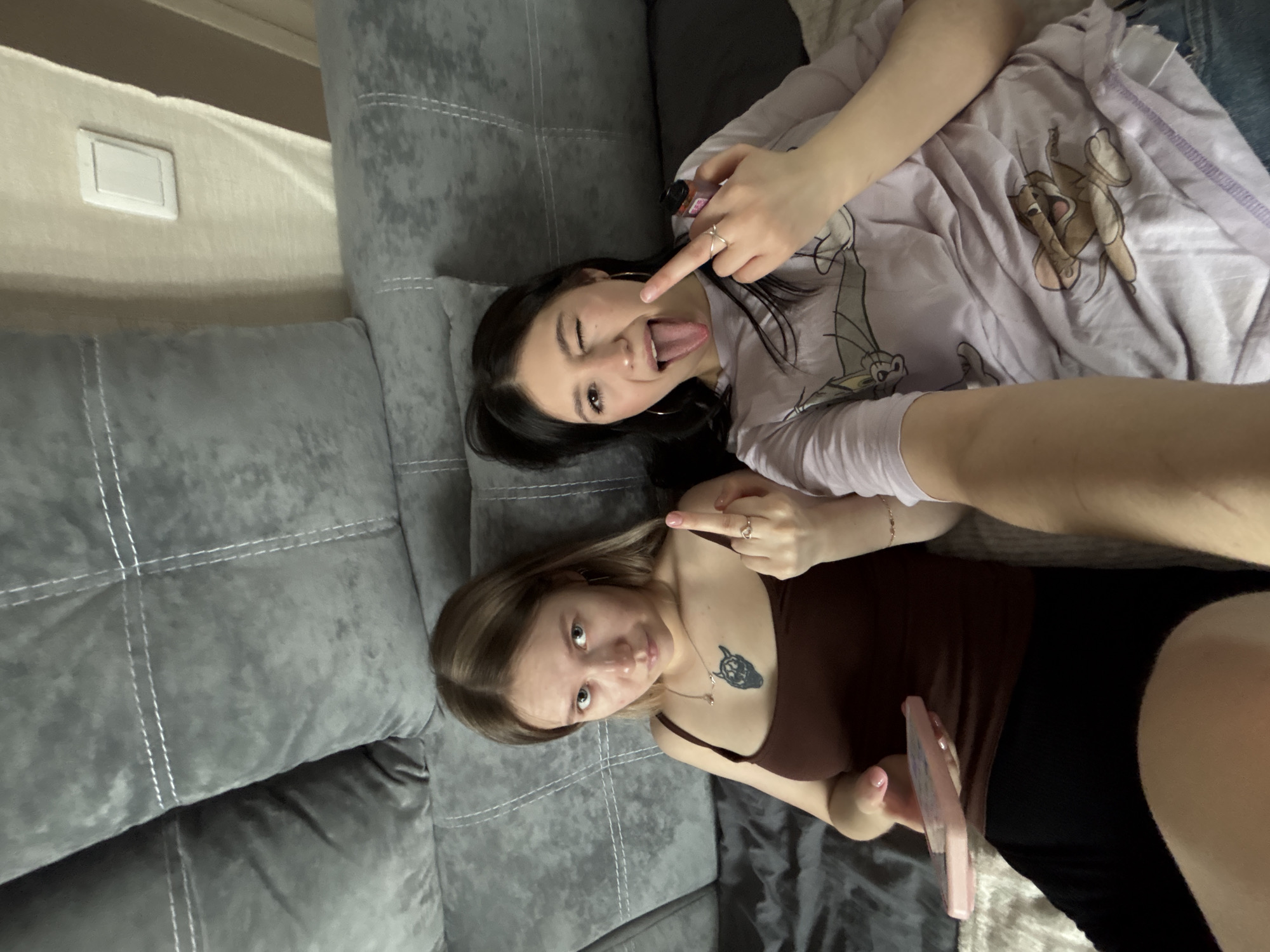 This image depicts two young adult females indoors, seated on a grey upholstered sofa in what appears to be a private residence in Chaikovsky, Russia. The scene is captured from a high-angle perspective, consistent with a selfie.
The female on the left has light brown hair and is wearing a dark brown tank top and black bottoms. She exhibits a dark-colored tattoo on her upper chest, appearing as a stylized animal head. A necklace with a pendant is visible around her neck. Her right hand is raised, displaying an extended middle finger, while her left hand holds a light pink smartphone. Her gaze is directed towards the camera with a neutral to slightly stern expression.
The female on the right has dark hair and is wearing a light purple t-shirt featuring a "Tom and Jerry" cartoon character (Jerry). Her right eye is winking, and her tongue is extended. Her left hand holds a small, rectangular object, possibly an electronic vape device, which has a red and black design. Her right hand is also raised, displaying an extended middle finger.
Both individuals wear rings on their right hands. They are seated on a light-colored blanket or throw covering the sofa seat. The background consists of multiple grey sofa cushions with white stitching, and a light-colored wall with a white electrical light switch visible in the upper right quadrant.