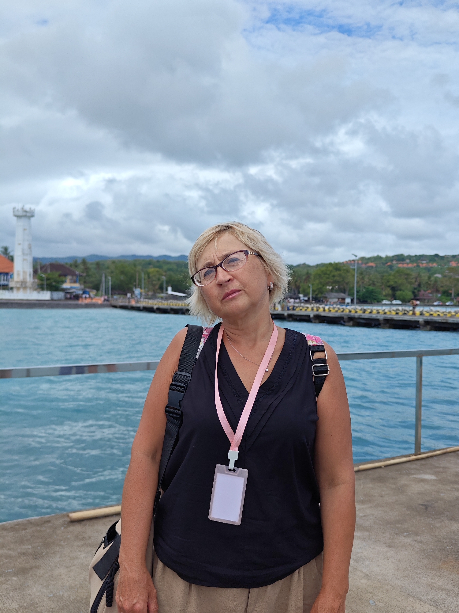 A woman stands on a pier in Kampung Toyapakeh, Indonesia, with the ocean in the background under a cloudy sky. She wears glasses, a black tank top, and a khaki skirt, with a pink lanyard around her neck. A backpack is slung over her shoulder. In the background, a white lighthouse is visible on the left, and a pier extends into the water on the right, with land and trees in the distance. A metal railing runs along the edge of the pier where the woman stands.
