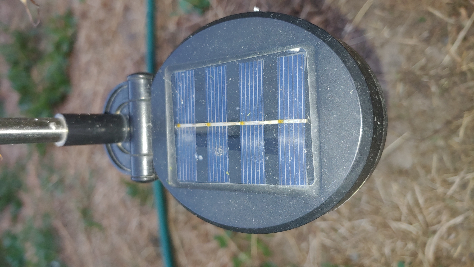 The image shows a close-up, top-down view of a solar-powered garden light. The light has a circular, black casing with a visible solar panel on its surface. The solar panel is blue with thin, horizontal lines representing its photovoltaic cells, bisected by a vertical strip with small yellow marks. The light is mounted on a thin, metallic pole, which is mostly out of focus at the top of the frame. The background is blurred, depicting dry grass and some green foliage, suggesting an outdoor, possibly garden or yard setting. A thin green hose is visible in the upper left corner of the image. No people or animals are present, and there is no discernible indication of the specific location within the city of Mukachevo, Ukraine. The scene is static and appears to be taken during daylight.