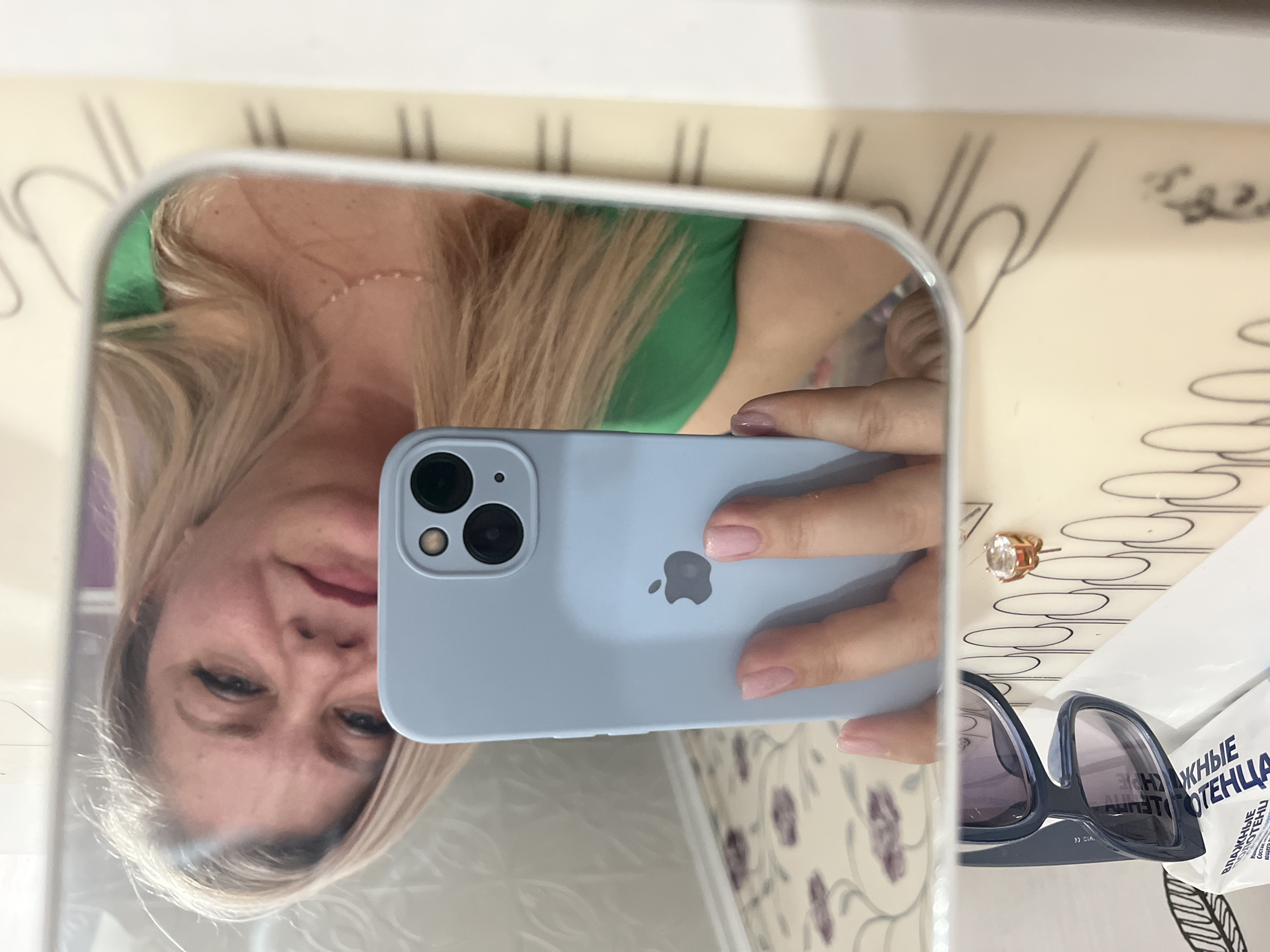 A mirror selfie shows a woman holding an iPhone 13 in a light blue case. The woman has blonde hair and is wearing a green top. The background includes sunglasses, an earring, and a package of wet wipes with Russian text. The environment appears to be indoors, potentially a bathroom. The location of Izhevsk, Russia is not discernible from the image.