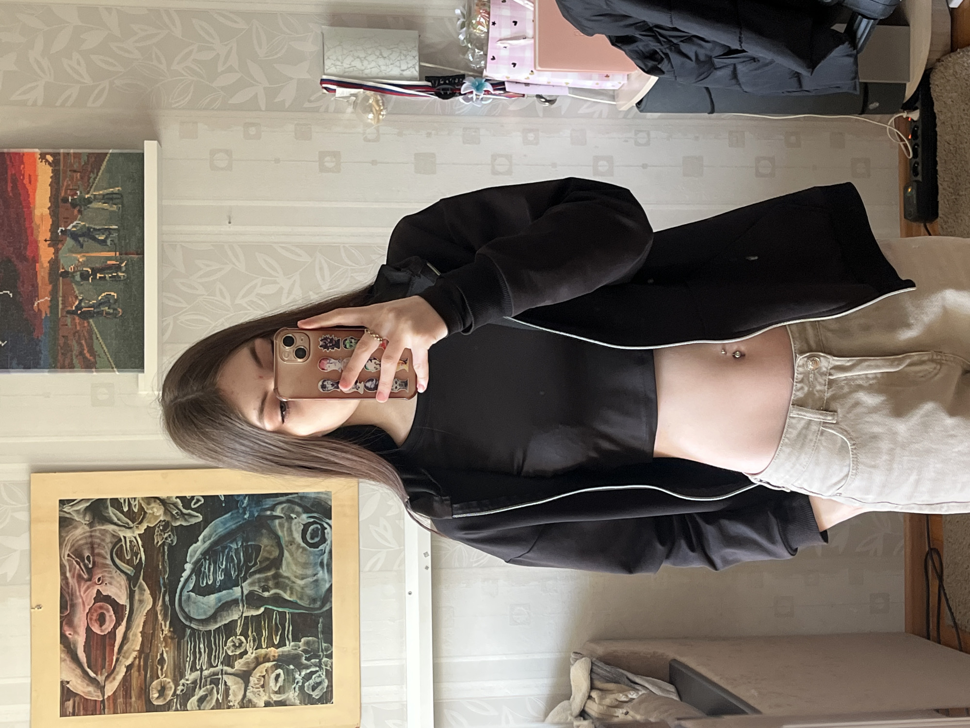 A young woman is taking a selfie in what appears to be a bedroom. She is wearing a black top and jacket paired with beige pants, revealing a navel piercing. Her phone has stickers on the back. The background features patterned wallpaper, two framed artworks on the wall, and a collection of items on a shelf above, including a pink notebook and some miscellaneous objects. An electrical outlet strip is visible in the corner. The location cannot be identified as Saint Petersburg, Russia based on the visual information provided.