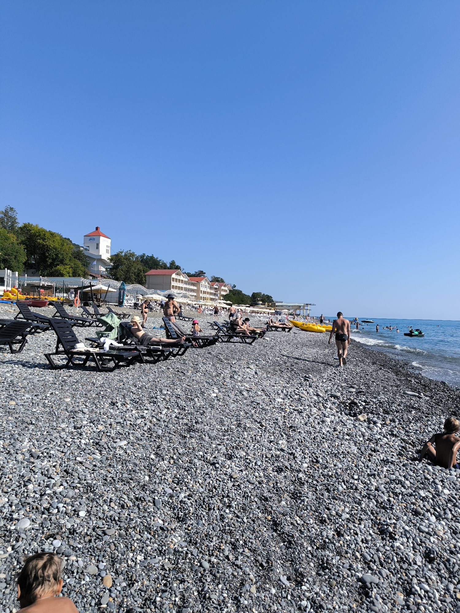 The image depicts a crowded pebble beach in Sochi, Russia, on a clear, sunny day. The foreground is dominated by dark grey and black pebbles. In the mid-ground, rows of empty black sun loungers are set up, with a few people sunbathing on some. Further back, more people are visible on the beach and near the water. A man in black swim trunks walks away from the camera towards the sea.
Beyond the beach, there is a line of buildings with red-tiled roofs, possibly hotels or apartment complexes, backed by lush green trees on a hillside. A tall white building with a red roof and the word "RIVIERA" is prominent on the left side of the frame, indicating a specific resort or establishment. The Black Sea stretches out to the horizon, with small waves lapping at the shore. Several yellow kayaks and a green jet ski are visible on the water, along with other people enjoying water activities. The sky is a brilliant, cloudless blue. In the far bottom left corner, a person's back and head are visible, looking out towards the beach. In the lower right corner, a person is sitting on the pebbles, facing away from the camera.