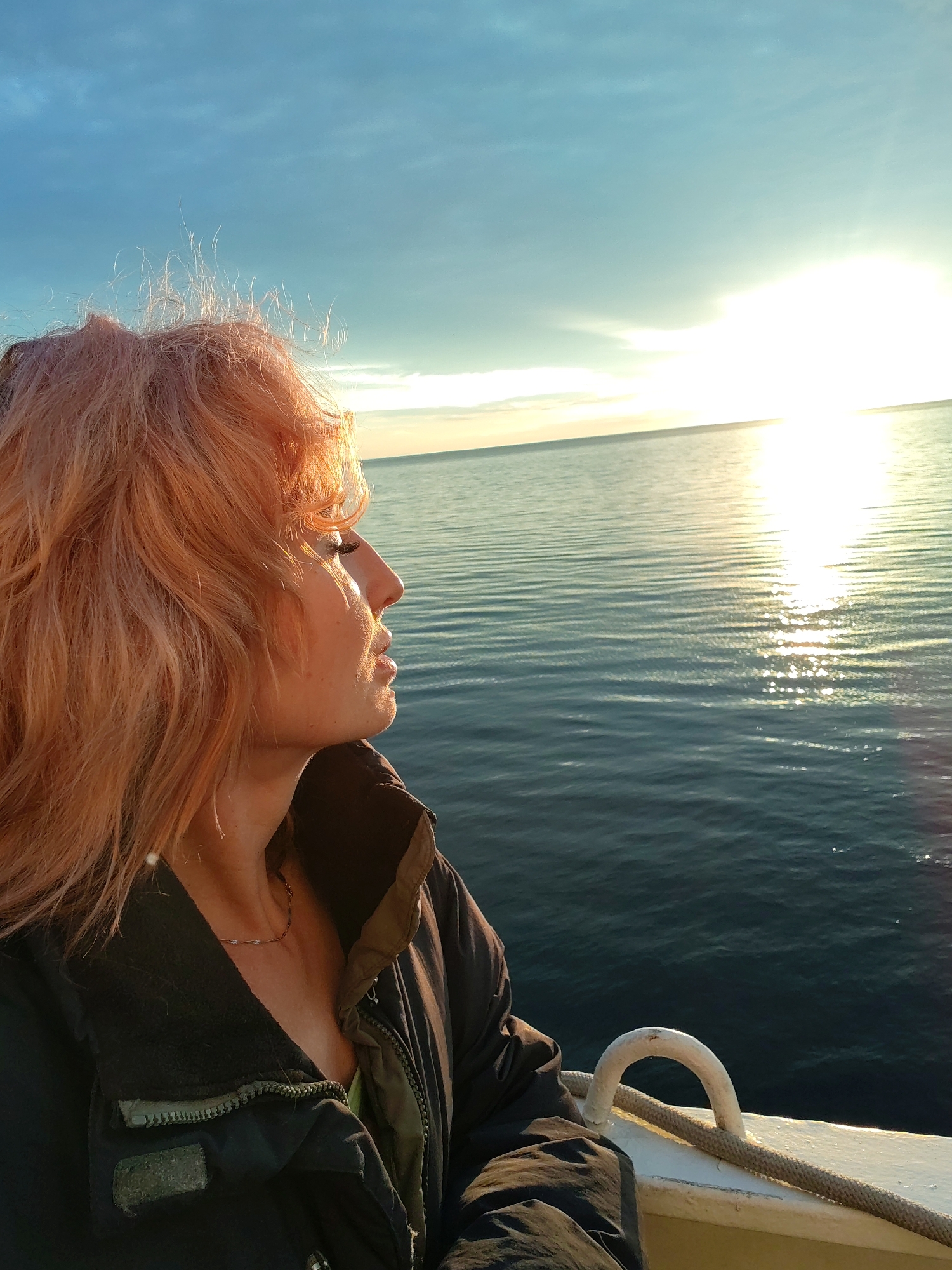 A woman with reddish-pink hair is seen in profile, looking out at a calm body of water at sunset. The sun is low on the horizon, casting a bright reflection on the water's surface. She is wearing a dark jacket with green accents and a necklace. The scene appears to be on a boat, with a white railing and a thick rope visible in the foreground. The sky is a clear blue with some wispy clouds. The image does not provide any specific visual cues to determine the exact location within the city of Shcheleiki, Russia.