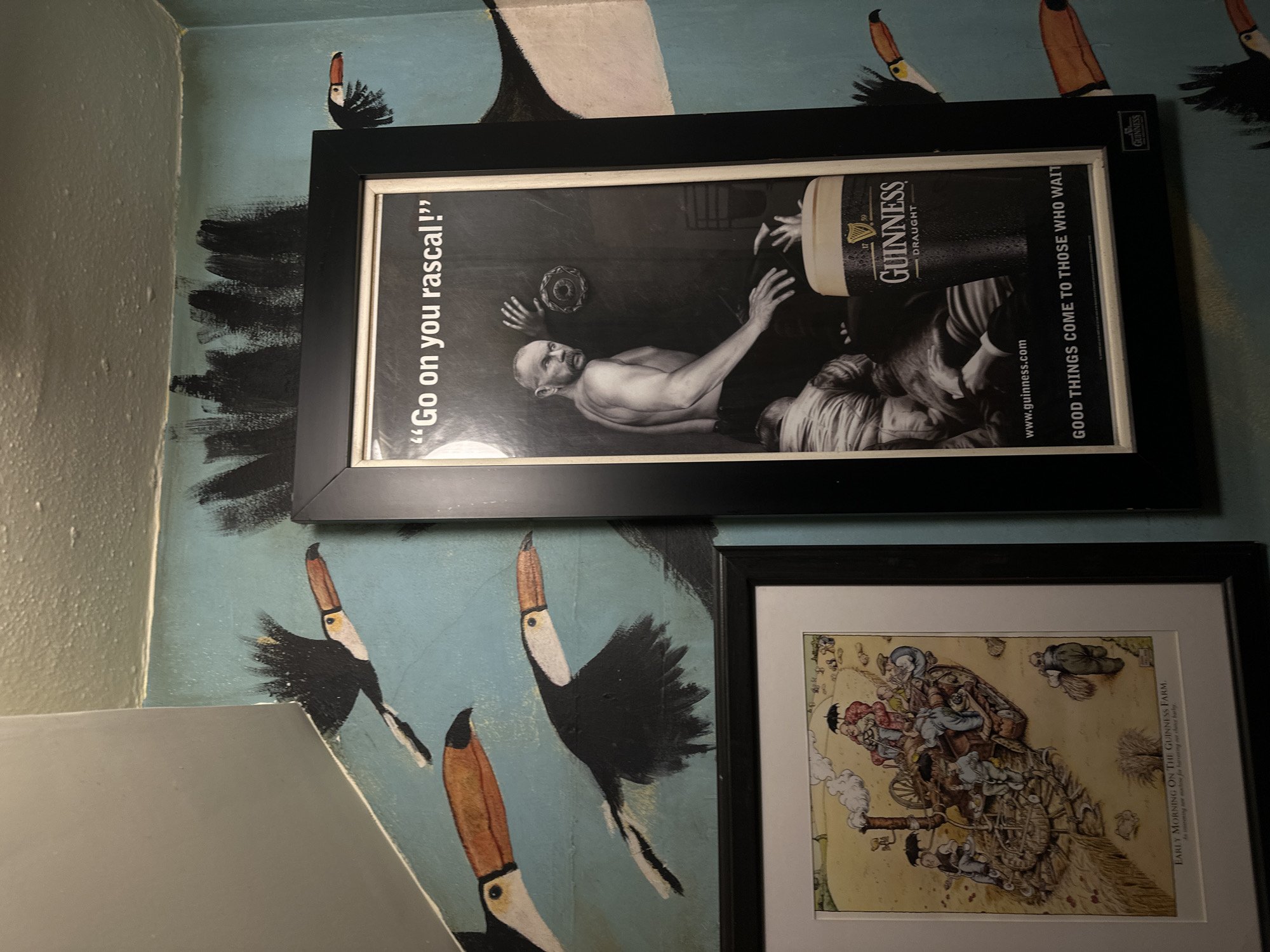 The image depicts a wall adorned with two framed pictures. The top picture is an advertisement for Guinness, featuring a monochrome image of a man with his shirt off, alongside a pint of Guinness, the slogan "Go on you rascal!", and the tagline "Good things come to those who wait". The lower picture, also framed, shows a complex, cartoon-like drawing titled "Early Morning on the Guinness Farm." The wall itself is painted light blue, decorated with illustrations of black and orange toucans. The location seems to be an interior, potentially a pub or a home. There is no explicit reference to the city of London in this image.