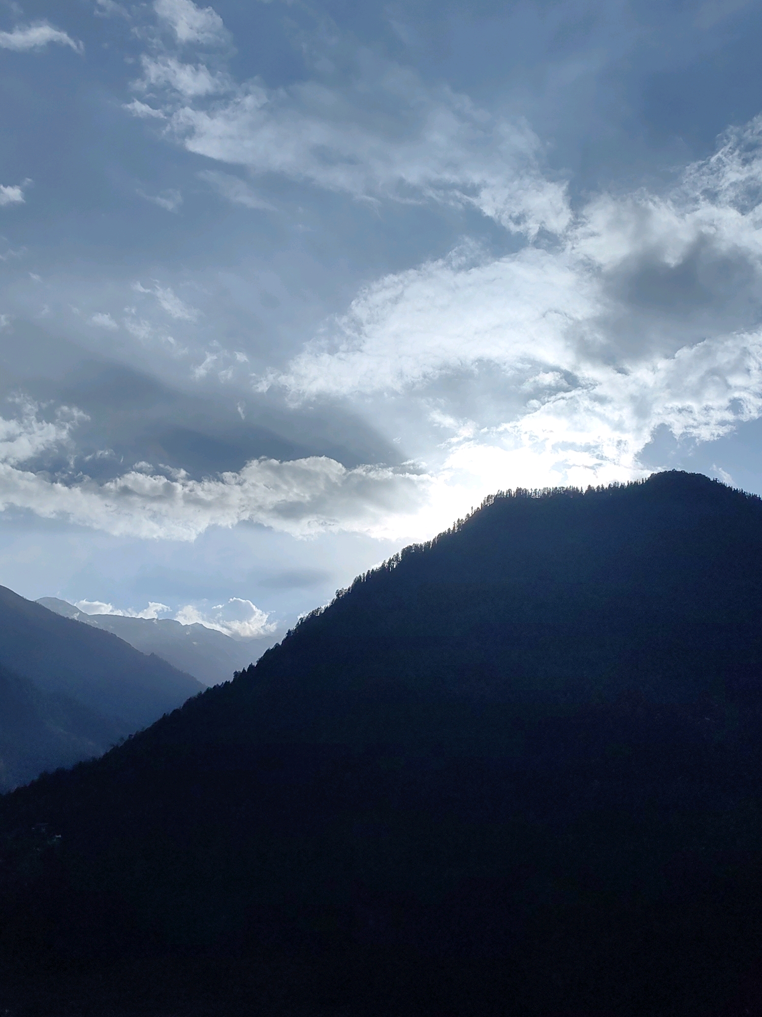 The image captures a dramatic mountain landscape at dusk, likely near Vashisht, India. The foreground is dominated by the dark, silhouetted shape of a mountain ridge, its peak thinly outlined by the fading sunlight and dotted with the silhouettes of evergreen trees. Layers of receding mountains are visible in the background, fading into a hazy blue atmosphere. The sky above is a dynamic canvas of clouds, with patches of bright sunlight breaking through, casting ethereal rays. Some clouds are dark and heavy, suggesting recent or impending rain, while others are lighter and wispy, tinged with the warm hues of sunset. The overall mood is one of serene, yet powerful natural beauty, with the interplay of light and shadow creating a sense of depth and mystery. There are no people or man-made structures clearly visible in the frame, emphasizing the wild and remote character of the location.