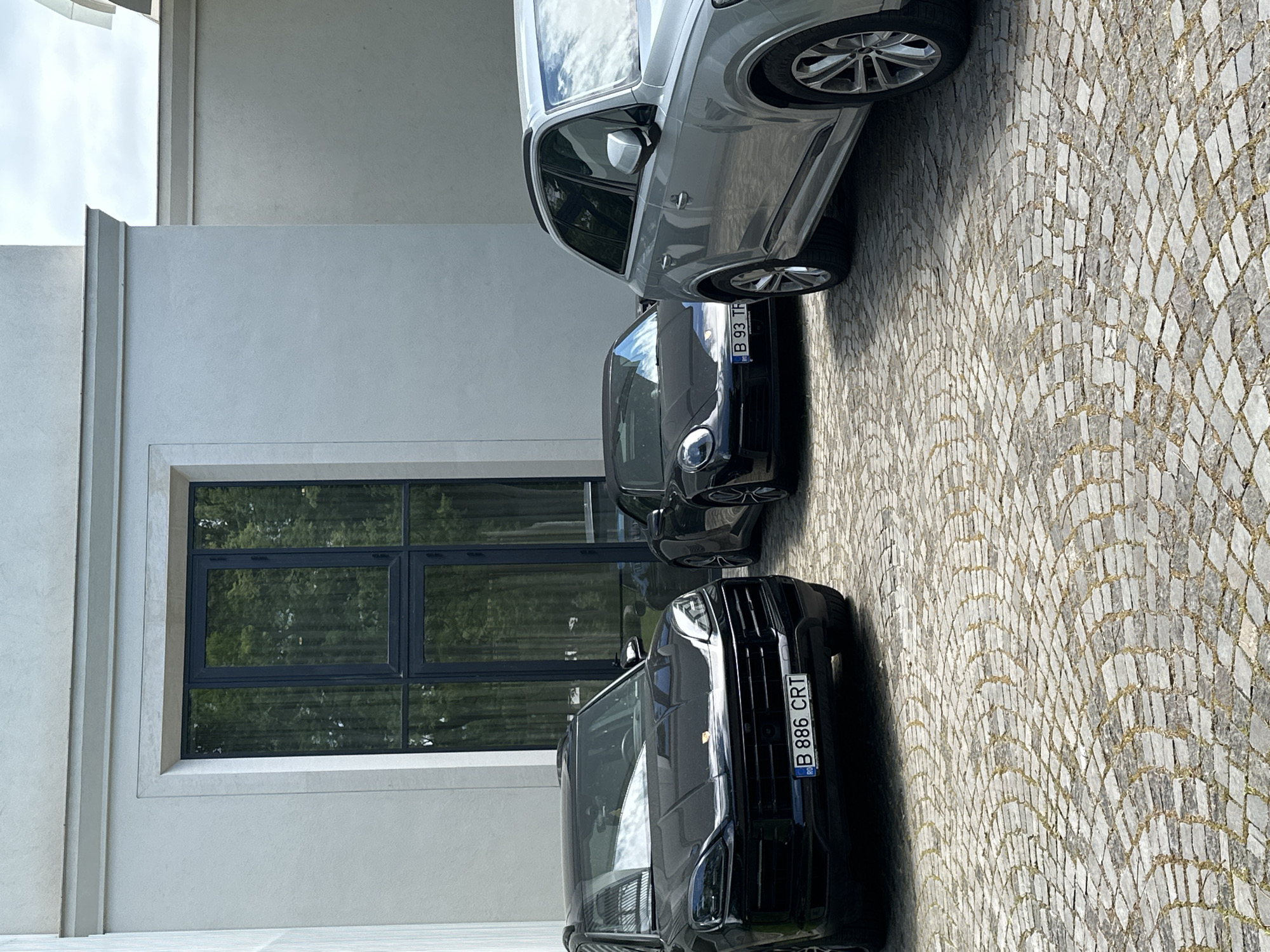 The image shows three parked cars on a cobblestone surface next to a building with a window. The cars are parked in a line. The first car is black with a license plate that reads "B 886 CRT". The second car is also black with a license plate that reads "B 93 TFF". The third car is silver. The window reflects trees. The location cannot be determined as Varna, Bulgaria.