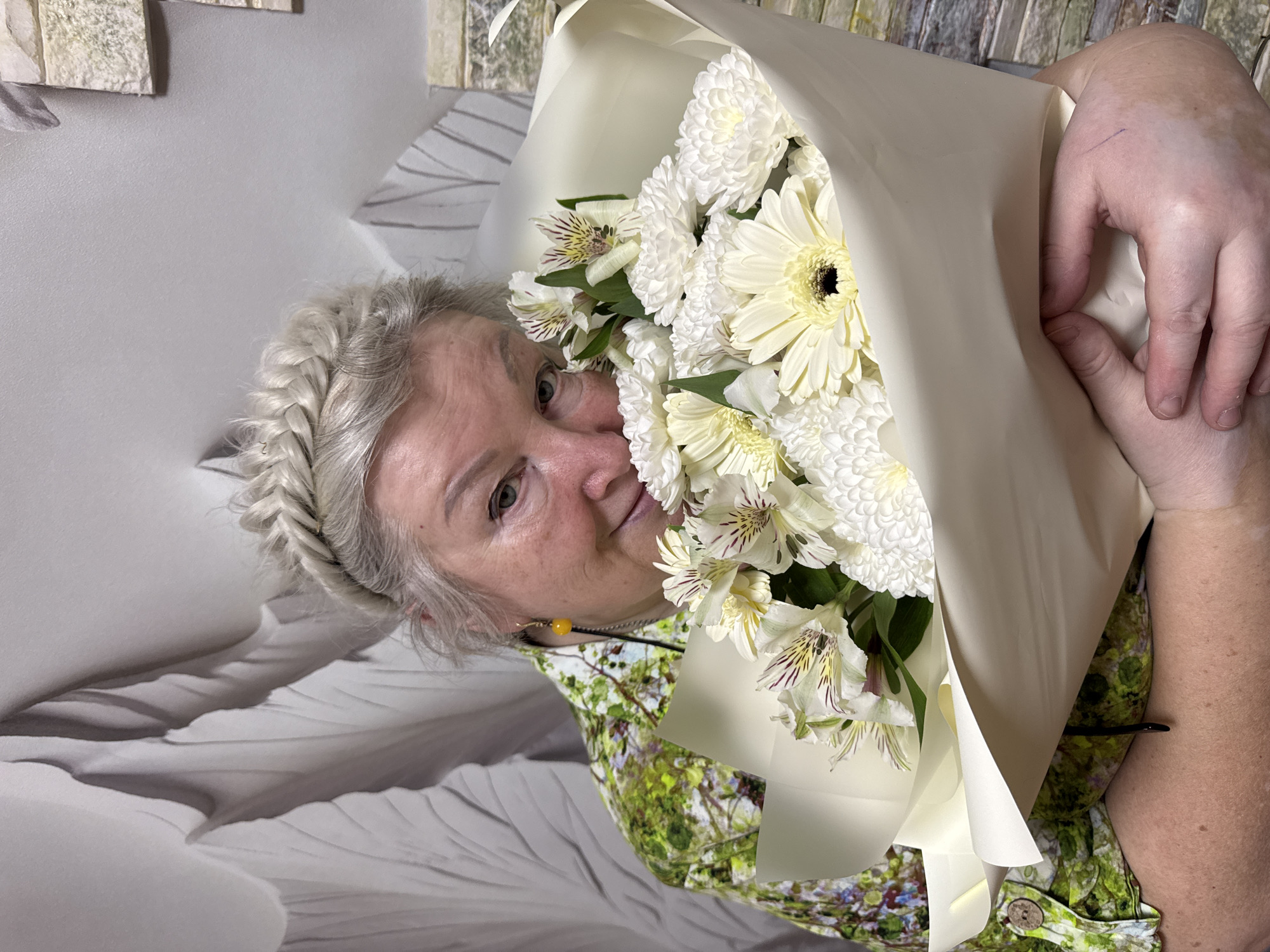 A woman with a gray braid is holding a bouquet of white flowers in a light-colored wrapper. She is wearing a green and white patterned shirt. The background appears to be a wall with a patterned design. The photo was taken indoors, likely in Chaikovsky, Russia.