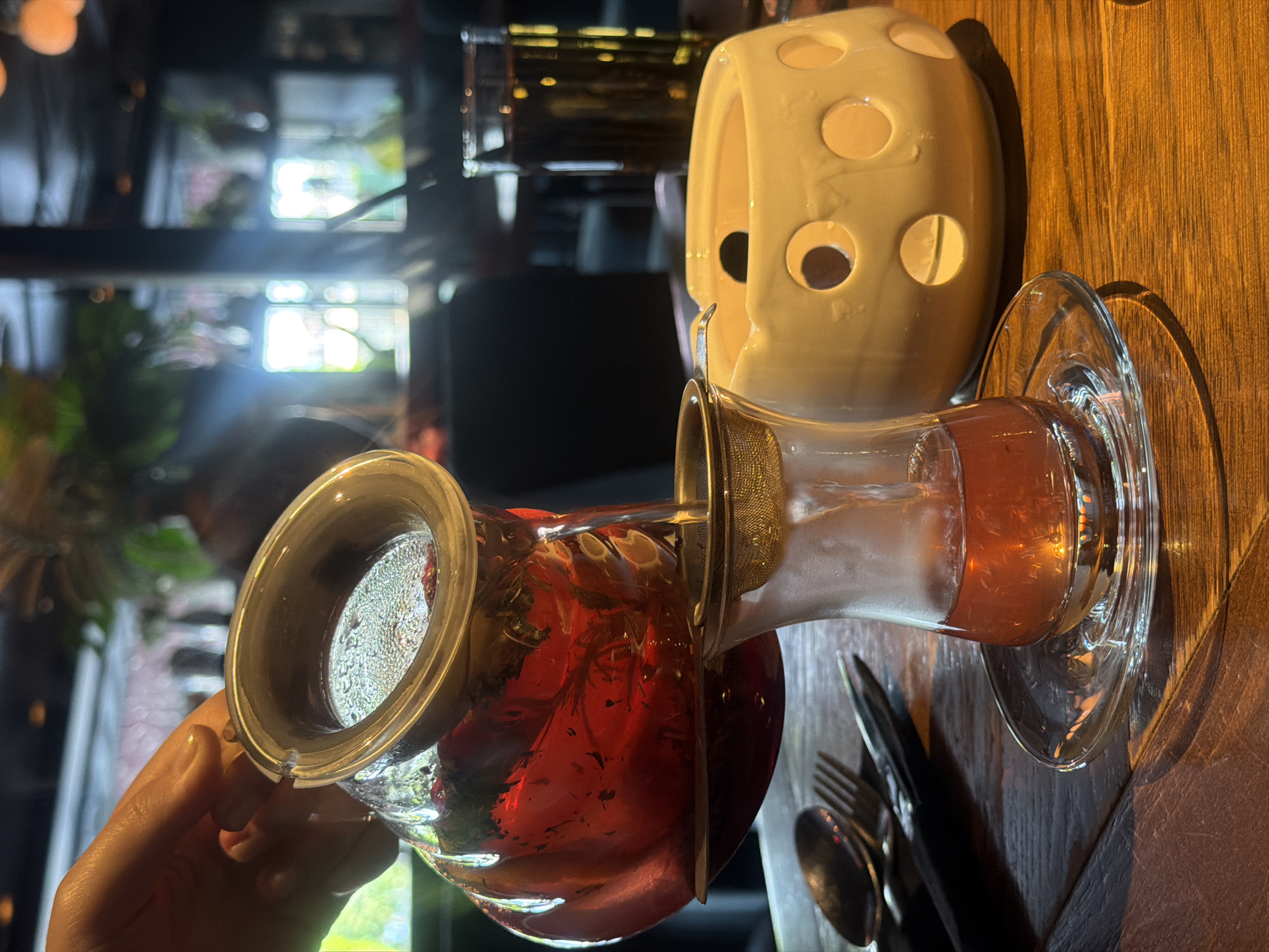 The image shows a hand pouring a red liquid from a glass container with a strainer into another glass container. The container sits on a glass saucer and the liquid appears to be tea. Behind the second container is a ceramic object with circular holes, reminiscent of Swiss cheese. Cutlery sits next to the saucer. The setting appears to be indoors, possibly a restaurant, given the lighting and table setting. It's not possible to confirm the specific location in Rostov-on-Don, Russia, from this image alone.