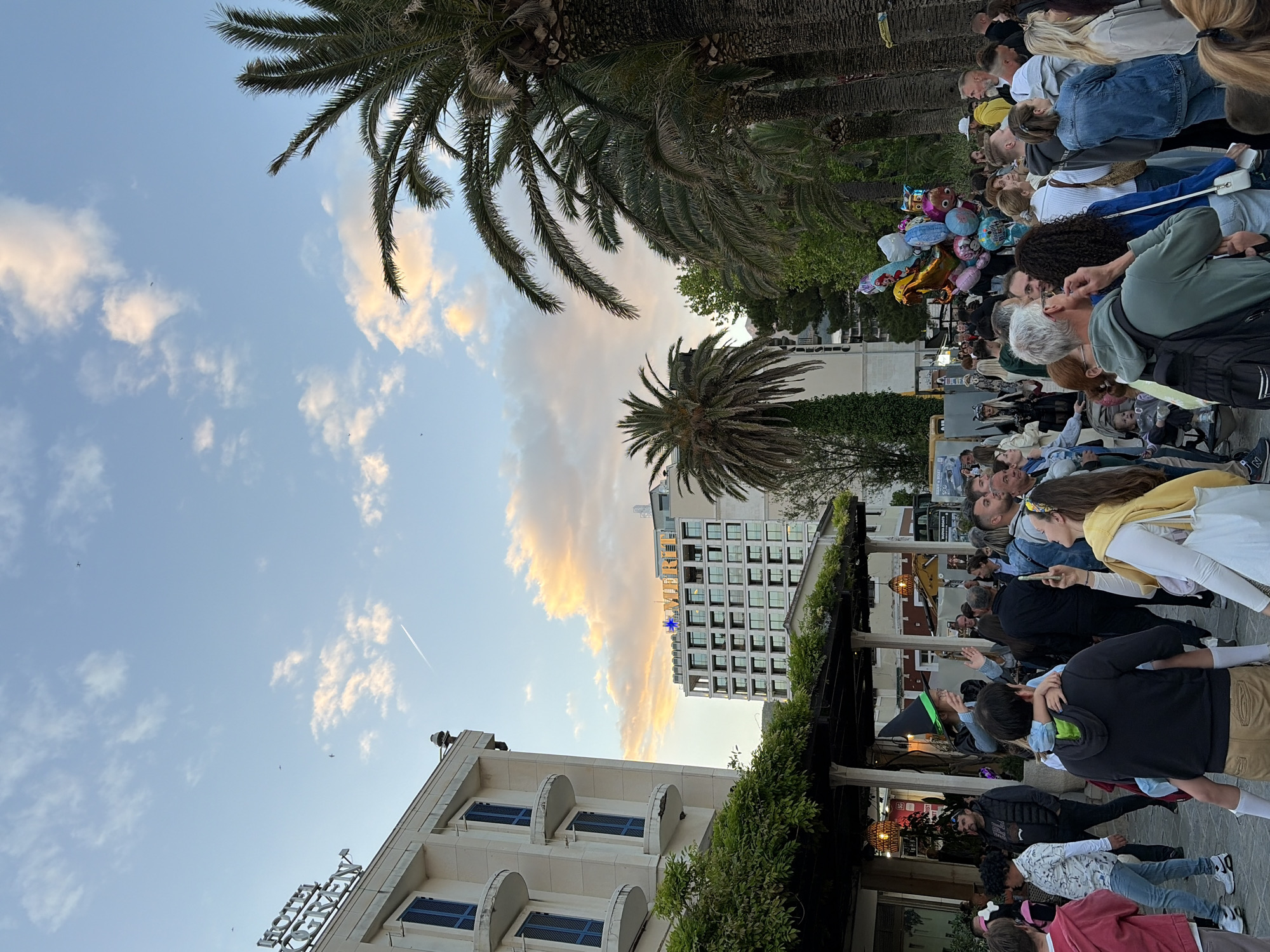 The image captures an outdoor scene in Budva, Montenegro. A dense crowd of people lines a street, possibly gathered for an event or parade. Many are looking towards a central point of interest. Several people are taking pictures with their phones.
Buildings are visible along the street, with palm trees lining the area. A building with white walls and blue window frames is seen, along with a taller building with a star logo on it. The sky is partly cloudy with light orange and yellow hues suggesting it might be near sunset.