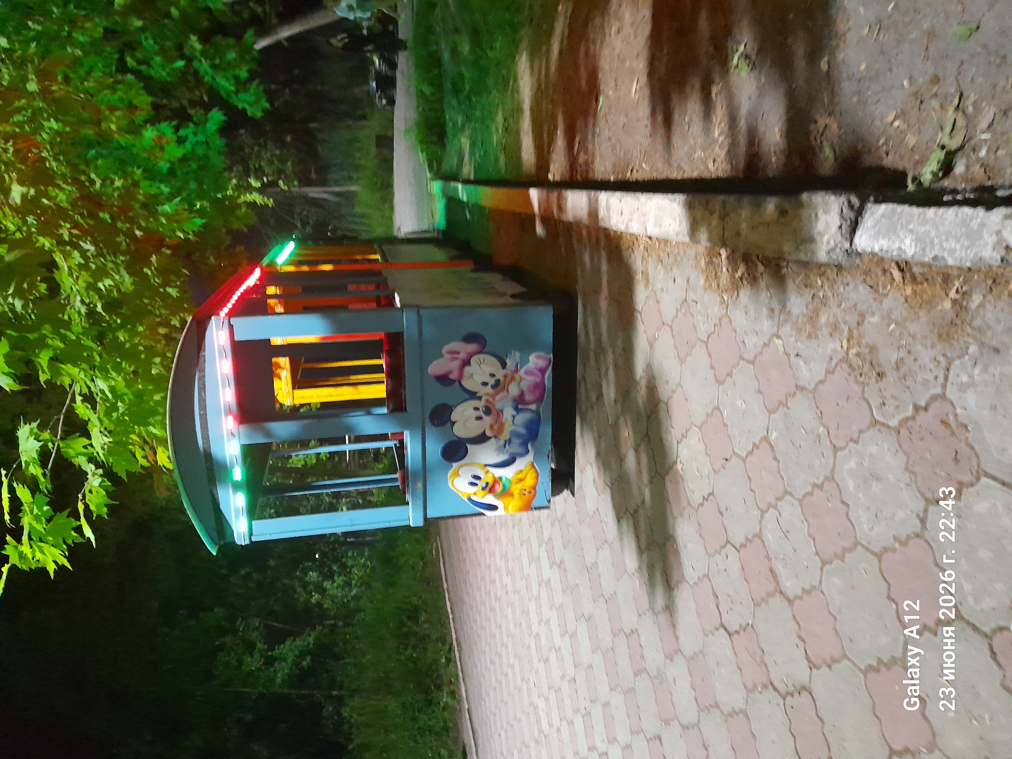 A blue kiddie train ride vehicle with cartoon characters Pluto, Mickey Mouse, and Minnie Mouse painted on its side is parked on a paved walkway at night. The vehicle has colorful LED lights along its roof. The walkway is made of interlocking hexagonal pavers in shades of pink and gray. Green trees with bright green foliage are visible in the background. The scene appears to be in a park or recreational area. A concrete curb runs alongside the walkway. The timestamp "23 июня 2026 г. 22:43" and "Galaxy A12" are overlaid in the bottom left corner of the image.
