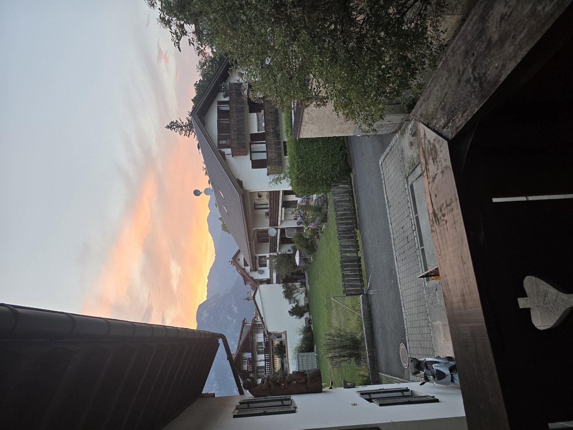 This is a medium shot, taken from a balcony or elevated vantage point, looking out at a residential street in Garmisch-Partenkirchen, Germany, during a sunset. In the foreground, a dark wooden balcony railing is visible, featuring a carved heart shape. Beyond the railing, a light-colored building is on the left, with dark shutters on a window. A light blue scooter is parked on a paved area in front of this building.
The scene opens up to a well-kept green lawn, bordered by a wooden fence. Several residential buildings with pitched roofs are situated across the street. These houses appear to be multi-story with balconies and windows. Trees and lush green hedges surround the properties. In the background, a mountain range is visible, silhouetted against a sky filled with soft orange, pink, and white clouds. The overall atmosphere suggests a tranquil evening in a European town.