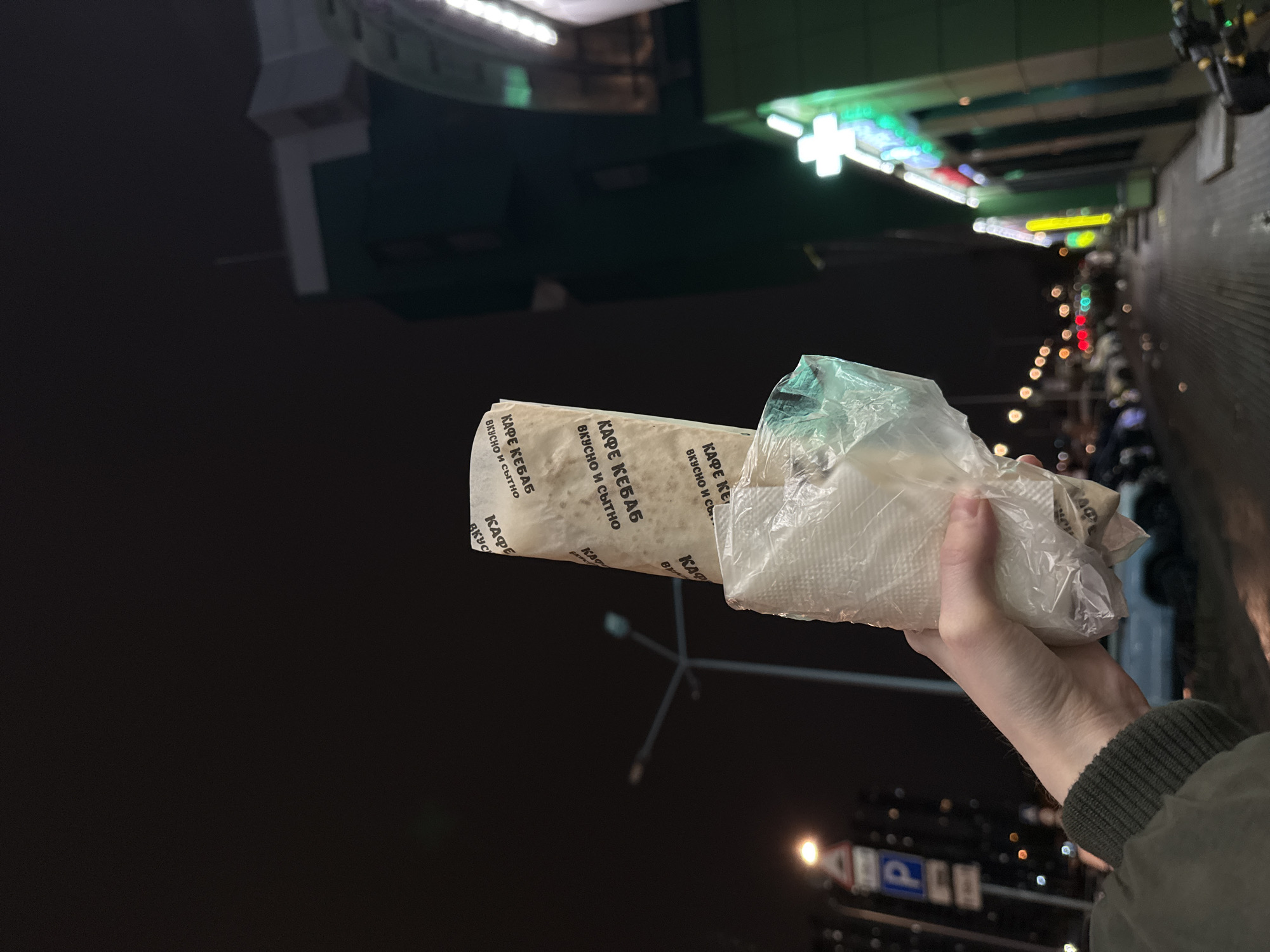 The image, correctly oriented, shows a person's right hand holding a wrapped meal against a blurred nighttime urban background. The food, likely a kebab or shawarma, is long and cylindrical, wrapped in white paper and partially covered by a clear plastic bag. The paper wrapper prominently features Cyrillic text, repeated multiple times, which reads "КАФЕ КЕБАБ" (KAFE KEBAB) and "ВКУСНО И СЫТНО" (DELICIOUS AND FILLING). The hand wears a sleeve of an olive green jacket with a ribbed cuff.
The setting is an illuminated city street at night, with a dark sky dominating the upper left. The background is out of focus, creating a bokeh effect from various artificial lights. Numerous colorful neon signs and building lights are visible, including a distinct bright green cross, often associated with a pharmacy. Other street lights and blurred signs, such as a blue sign with a white 'P' for parking, further indicate an urban environment. The scene suggests a casual moment, possibly after purchasing street food, on an evening walk in Saint Petersburg, Russia.
