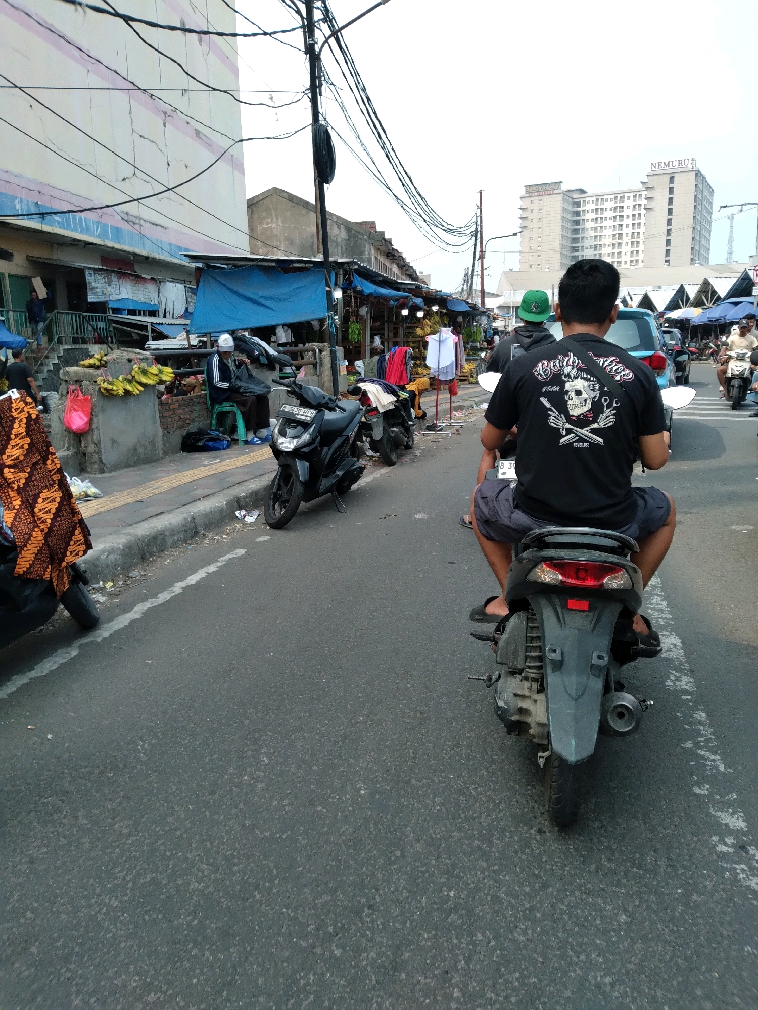 The image depicts a street scene in a commercial area of South Tangerang, Indonesia. In the foreground, a person is riding a motorcycle, seen from behind. They are wearing a black t-shirt with a graphic print of a skull and scissors, dark shorts, and black sandals. The motorcycle is dark gray and has a license plate with the letters "B" followed by numbers. The road is paved and shows some wear and tear, with a white line painted on the right side.
To the left of the road, there is a sidewalk and a market area. Stalls are set up with various goods, including bananas displayed on a raised concrete platform. Several motorcycles are parked along the sidewalk. People are seen browsing or working in the market. One person is sitting on a green stool, appearing to be working with fabric or clothing. Another person is visible in the background near a blue tarp.
In the middle ground, a light blue car is also on the road. Beyond the market, buildings of varying heights are visible. One prominent building in the background has the word "NEMURU" on its facade, a sign that appears to be for a hotel or residential complex. Overhead, a complex network of electrical wires crisscrosses the sky, supported by utility poles. The sky is overcast, suggesting potentially humid weather. The overall atmosphere is that of a busy, functional marketplace in an urban setting.