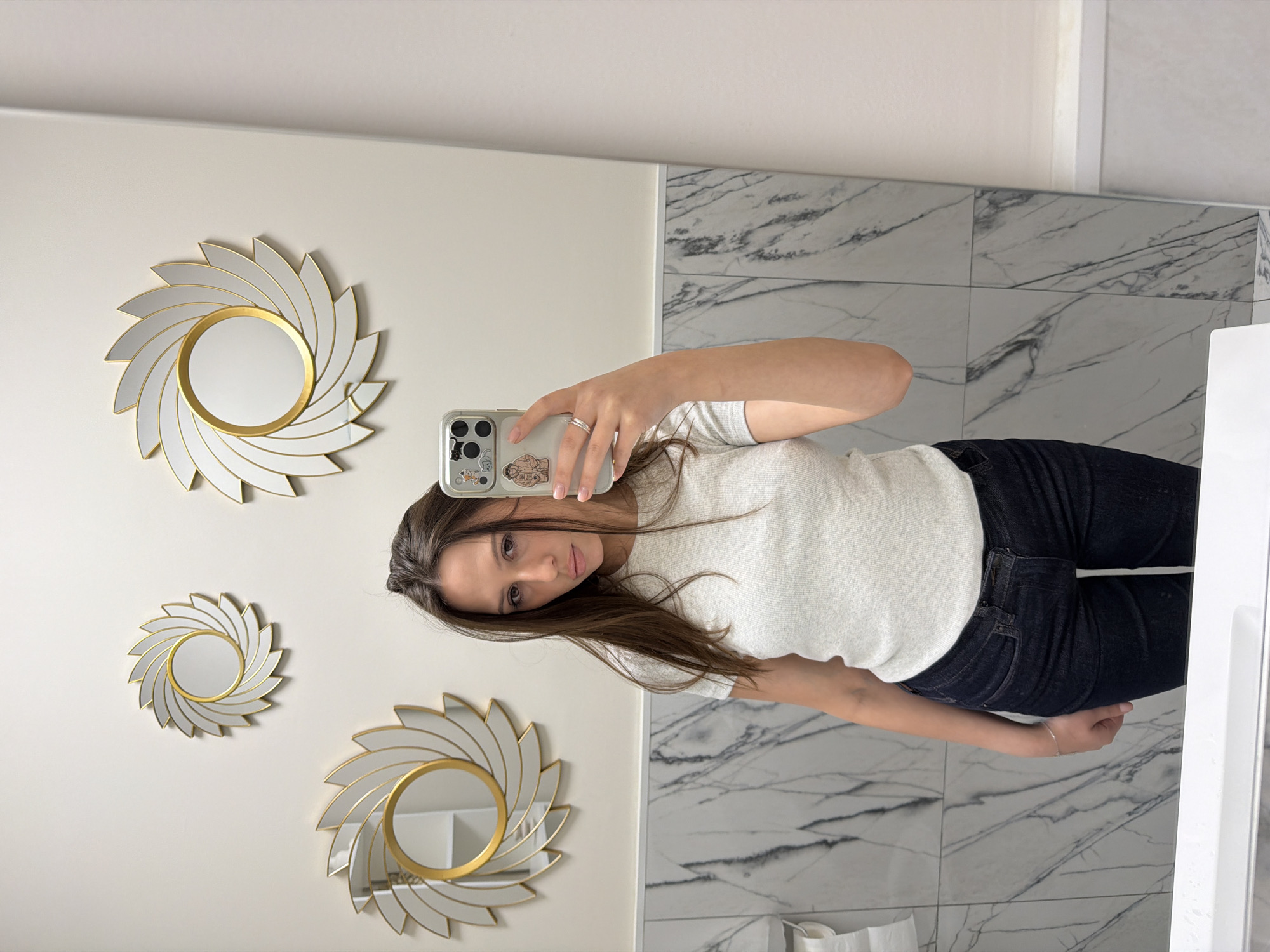 In a bathroom with marble-patterned tiled walls, a young woman takes a mirror selfie. She is wearing a light grey ribbed t-shirt and dark wash jeans. Her hair is long and brown, and she is wearing a ring on her left hand. Behind her, on the wall, are three decorative mirrors with gold frames, shaped like pinwheels or sunbursts. The lighting is bright, casting subtle shadows. The setting is a modern bathroom, likely in a residential or hotel property in Khimki, Russia.