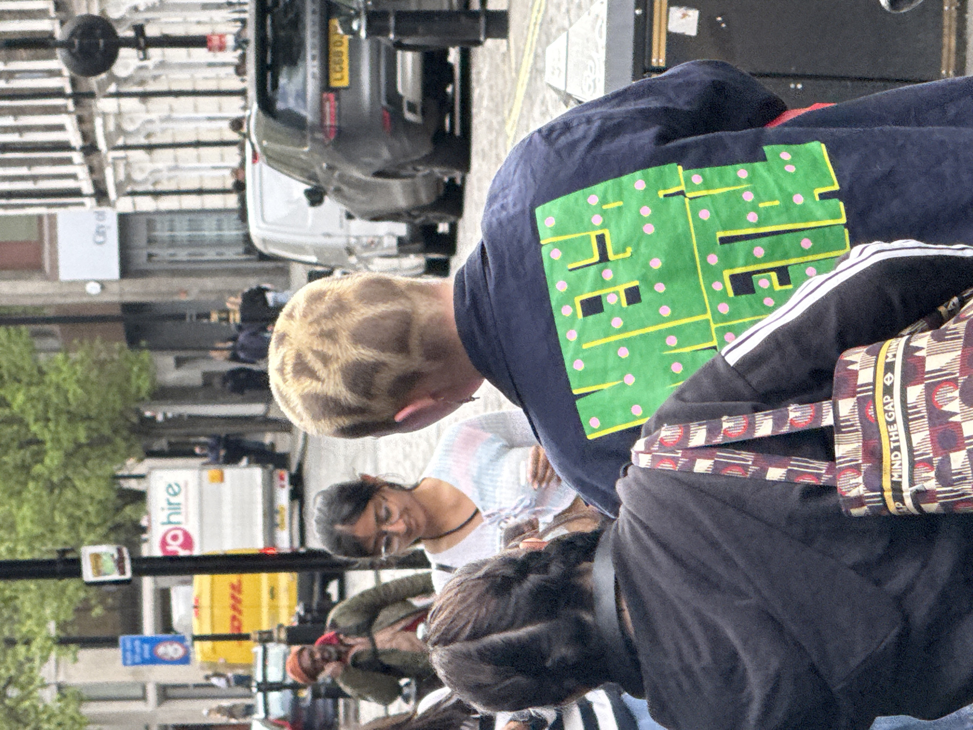 The image shows a group of people standing on a street in London, United Kingdom. A person with patterned short hair is wearing a navy blue shirt with a green printed circuit board design with pink dots. A woman wearing glasses is standing beside them, dressed in a light-colored patterned shirt. Another person with dark hair is wearing a dark jacket with white stripes on the shoulder and carrying a patterned bag with straps, which has the text "MIND THE GAP" printed on it. A yellow DHL van can be seen in the background, alongside other vehicles, including a YoHire van. A street sign is visible, attached to a lamppost.