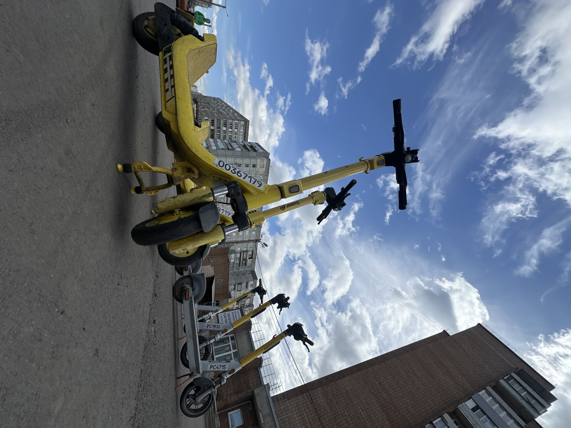 The image shows a row of parked scooters under a partly cloudy sky. The scooters are of two types: yellow and white. The yellow scooters are older models, while the white scooters are newer. One yellow scooter has the number "00367179" on it. The white scooters have numbers "PC1930" and "PC4715" visible. The scooters are parked on an asphalt surface, and there are buildings in the background. The buildings are of typical urban architecture. The scene is located in the city of Krasnoyarsk, Russia. There is a green figure at the top of the image, potentially a person.