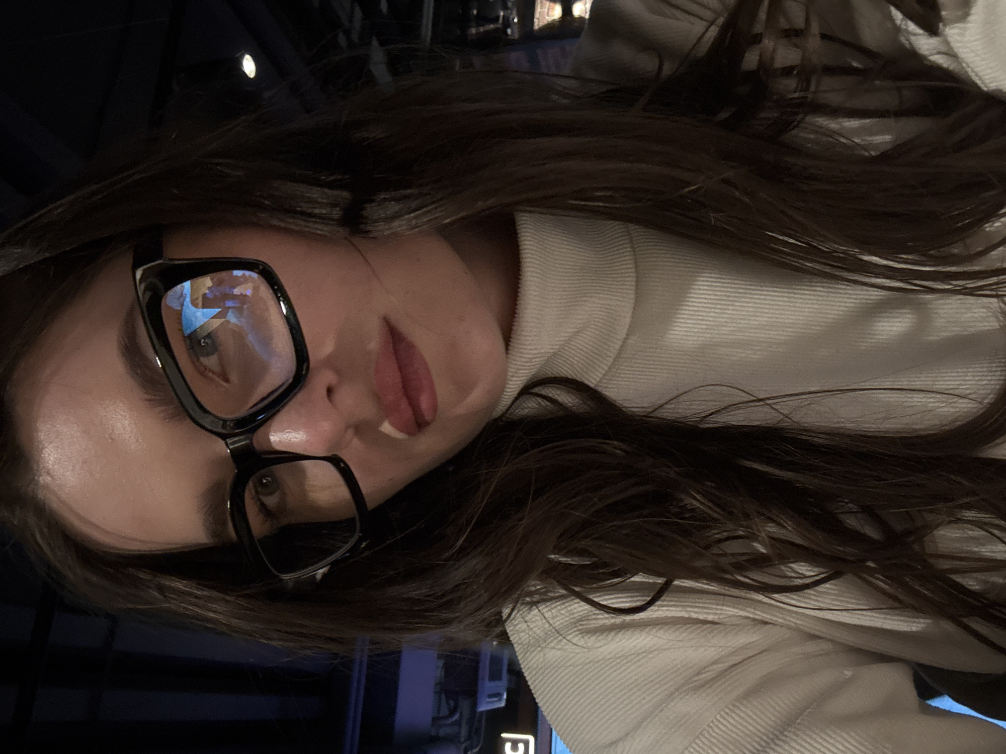 A young woman with long brown hair and wearing large black glasses and a white turtleneck sweater is in an indoor setting, possibly in Moscow, Russia. The reflection in her glasses suggests a building with blue elements, typical of urban Moscow architecture. The lighting suggests an evening or dimly lit interior.