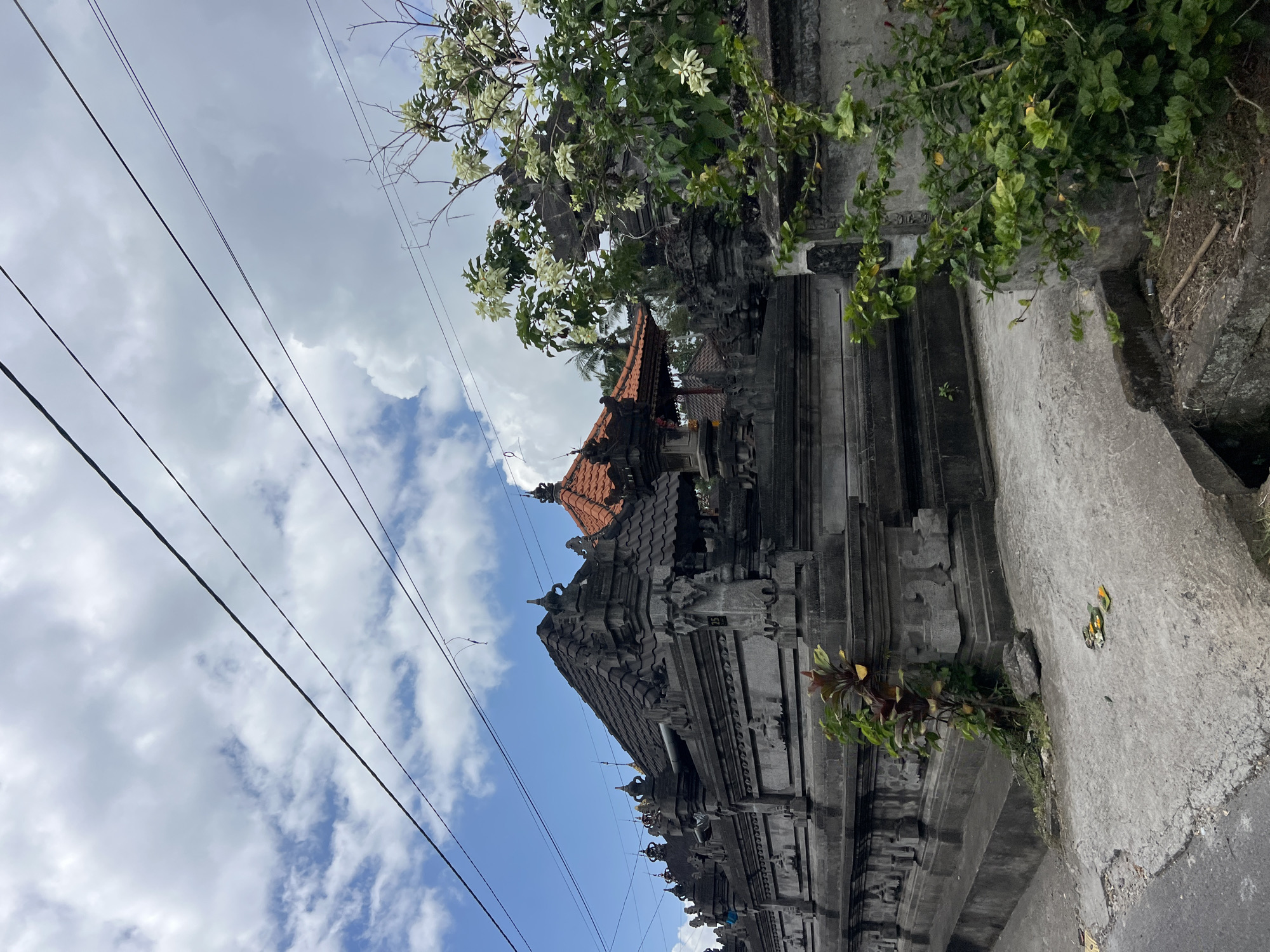 In Ubud District, Indonesia, a traditional Balinese temple complex is depicted under a cloudy sky. The temple features intricate stone carvings and tiered roofs, with one section topped with orange tiles. A lush green bush with white flowers grows in the foreground, obscuring part of the temple's facade. Power lines stretch across the sky, casting faint shadows. The ground in front of the temple is paved with concrete, showing some wear and tear, with a small offering of flowers and incense placed on the ground. A plant with reddish-green leaves grows from a crevice in the temple wall. The scene suggests a typical street view in a tropical urban environment.