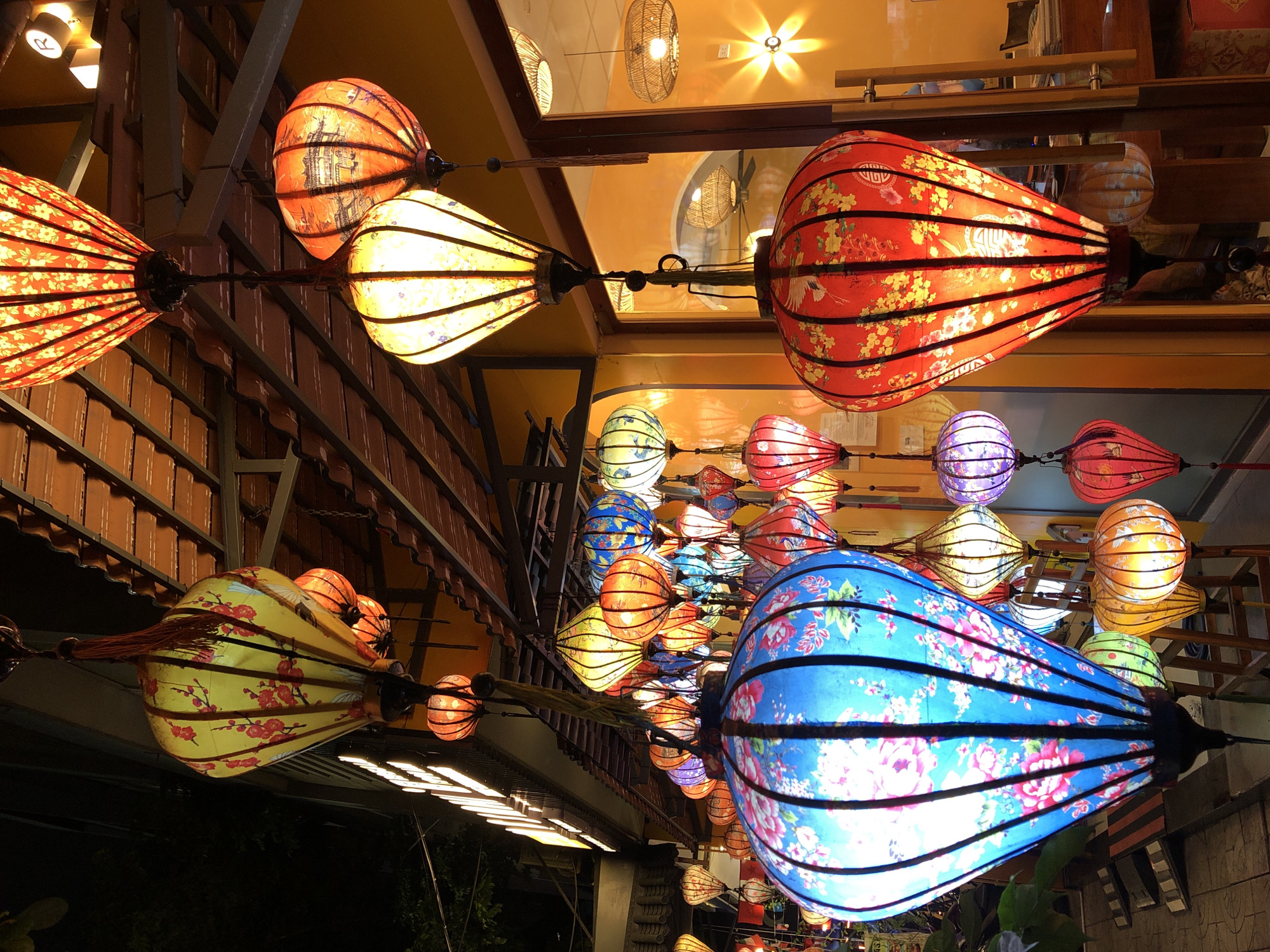 This image captures a vibrant nighttime scene on a street in Hoi An, a city in the Quảng Nam Province of Vietnam, renowned for its historic Old Town and its abundance of colorful silk lanterns. The foreground is dominated by a large, illuminated blue lantern decorated with pink flowers, hanging from a darker frame. To its right, a large red lantern, also illuminated, displays a pattern of yellow flowers and cranes. Behind these, a multitude of other lanterns in various sizes and colors—including yellow, orange, purple, and green—are suspended from above, creating a dazzling display. The lanterns hang along a covered walkway, with the roof structure visible overhead. The background shows the warm glow of interior lighting from shops or restaurants, with reflections visible in glass doors. The overall atmosphere is festive and inviting, characteristic of the evening ambiance in Hoi An's tourist areas.