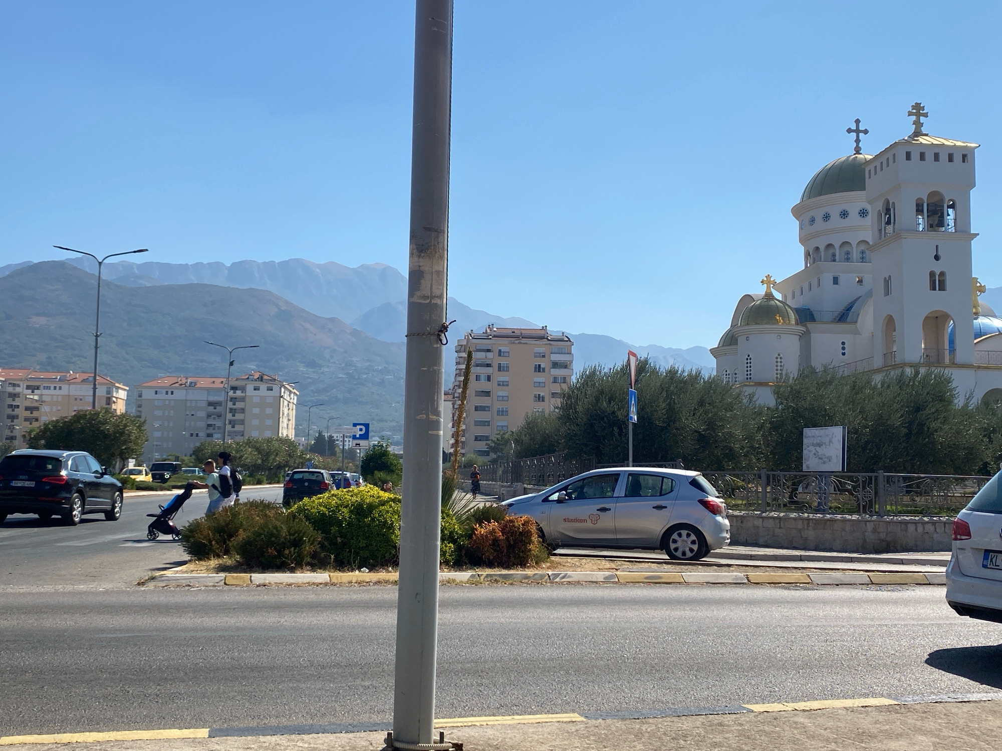 The image depicts a scene in the city of Bar, Montenegro, with a prominent white and gold Orthodox church in the background. The church features multiple domes and bell towers, characteristic of religious architecture in the region. In the foreground, a multi-lane road is visible, with several vehicles, including a black SUV, a silver hatchback, and the rear of a white hatchback. On the side of the road, there are pedestrians, including a person pushing a stroller, and a cyclist. Green trees and bushes line the roadside, separating the road from the buildings and the church. In the distance, mountains form a scenic backdrop under a clear blue sky. The overall impression is of a sunny day in a coastal city, with a mix of urban elements and natural landscapes.