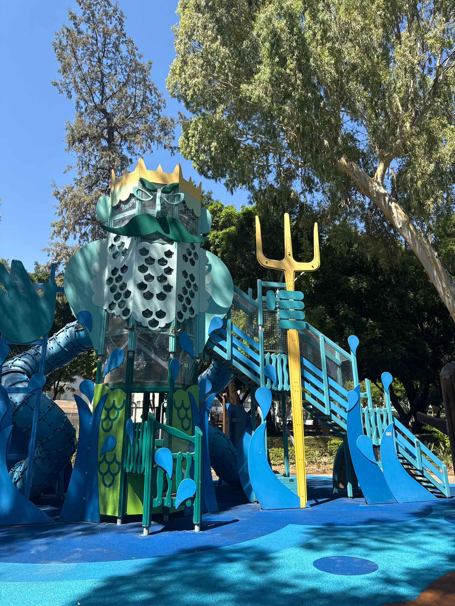 The image shows a playground structure at Limassol Municipality's Seaside Promenade in Cyprus. The playground is themed around a sea creature, possibly Poseidon, with a large blue and green head featuring a golden crown and glasses. There are slides and climbing structures in blue and yellow, designed to resemble waves and marine elements. A yellow trident is prominently displayed as part of the structure. The playground is situated outdoors with trees and blue sky visible in the background, and the ground is covered in a blue rubber safety surface. There are no people visible in the image.