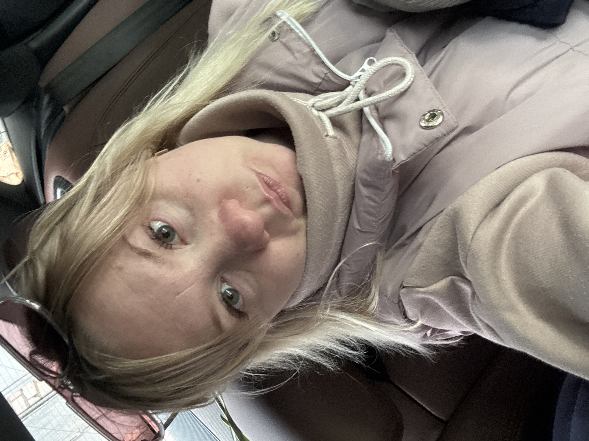 A woman with fair hair and light eyes is in a vehicle, possibly a car, based on the presence of a seat and what appears to be a window. She wears sunglasses on her head and a pink or light purple jacket with a hoodie. The interior of the car is brown. The specific location in the city of Chaikovsky, Russia, cannot be determined from the image.