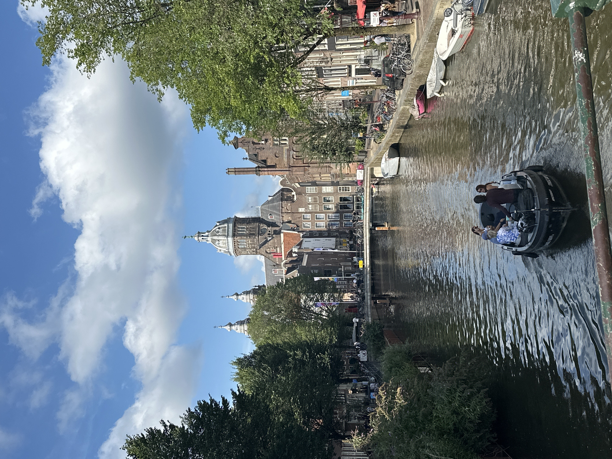 The image depicts a canal in Amsterdam, Netherlands, with a boat carrying four people navigating the water. In the background, an imposing building with domes and spires, likely a church or cathedral, is visible, flanked by characteristic Dutch architecture. Trees line the canal, and a bridge spans the waterway further in the distance. Several other boats are moored along the canal's edge, and bicycles are parked on the walkway. The sky is a clear blue with fluffy white clouds.