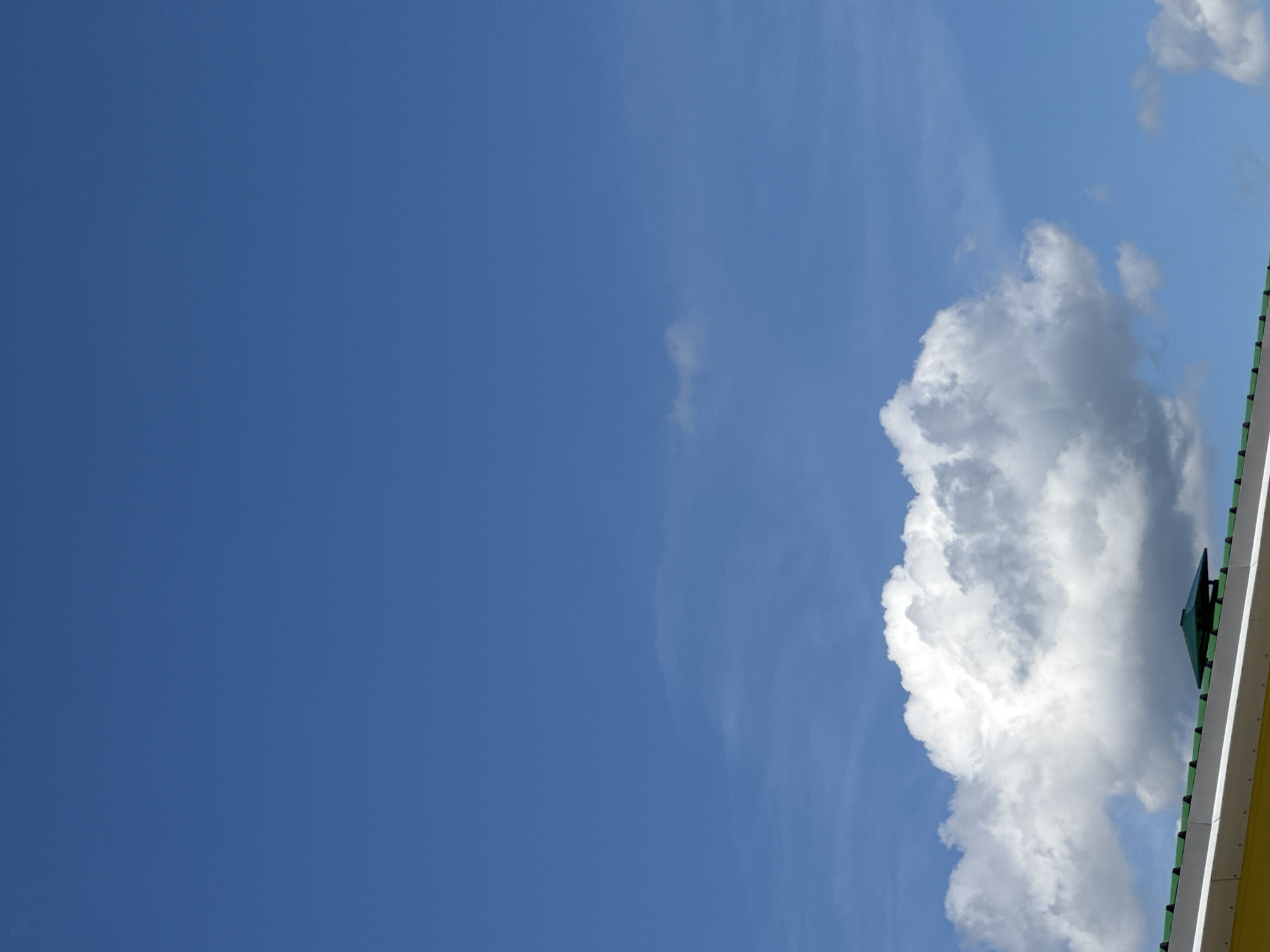 This is a vertical, eye-level shot of a bright blue sky with fluffy white clouds. A large cumulus cloud dominates the right side of the frame, appearing dense and textured with shaded areas suggesting depth. A sliver of a building's roofline is visible along the far right edge, featuring a yellow structure with a white trim and a dark green peaked element, possibly a small gable or ventilation. The building's edge is sharply defined against the softer forms of the sky.
The sky is a clear, vibrant blue, indicating a sunny day, likely midday or afternoon given the brightness and the nature of the clouds. The large cloud suggests fair weather, though its size and shading hint at the possibility of developing weather systems. The scene is outdoors, with the building indicating human construction within a natural landscape. There are no people or discernible activities taking place, only the interplay of sky, cloud, and a man-made structure. The overall mood is peaceful and serene, highlighting the vastness of the sky. There is no visible text in the image.