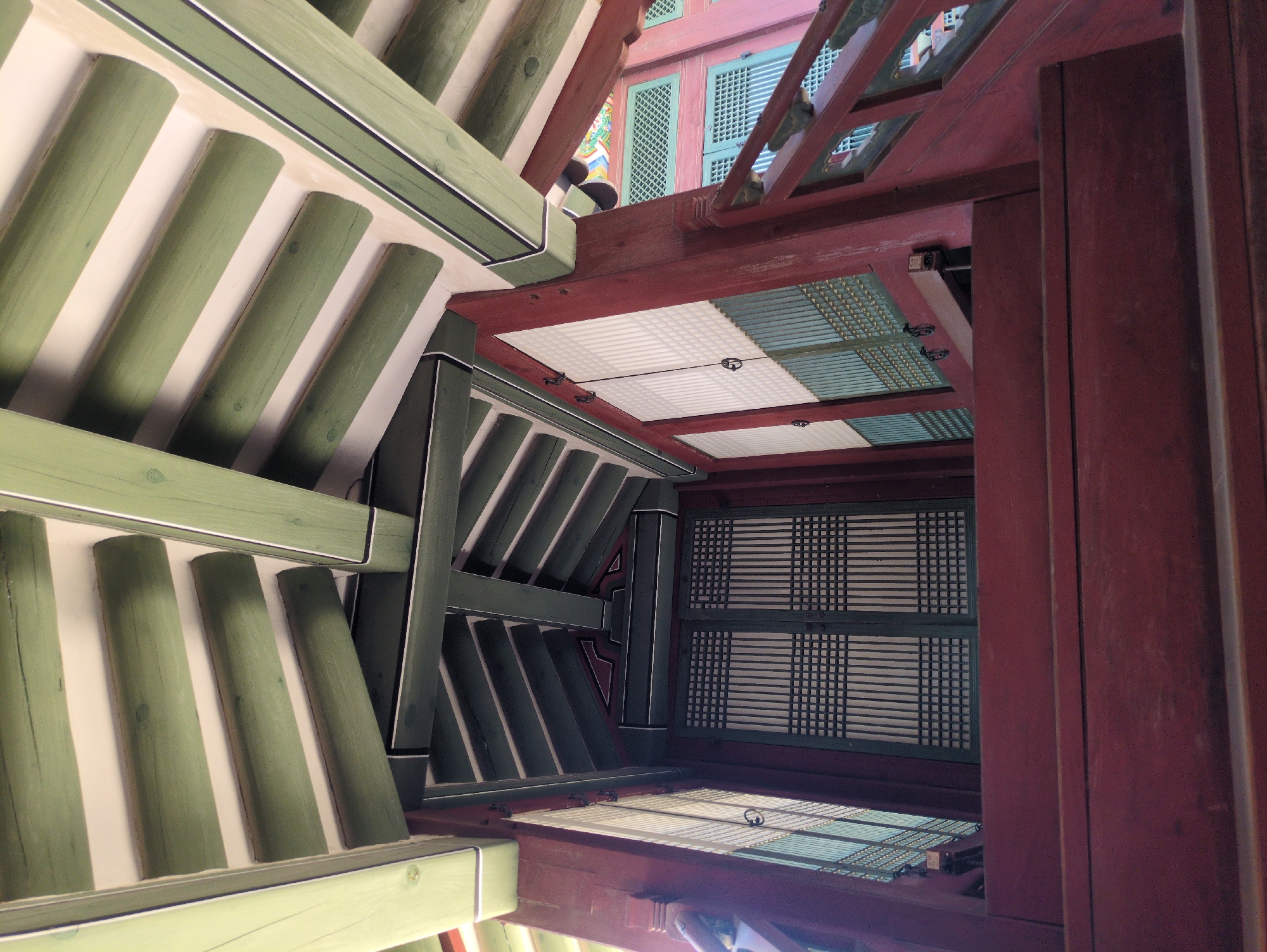 The image shows the interior architecture of a traditional Korean building, specifically a view looking upwards towards the roof and ceiling. The structure features wooden beams and supports, painted in muted green and deep red hues. The ceiling panels are constructed with a white lattice design. The image suggests the architecture is located in Seoul, South Korea, given the traditional Korean building styles.