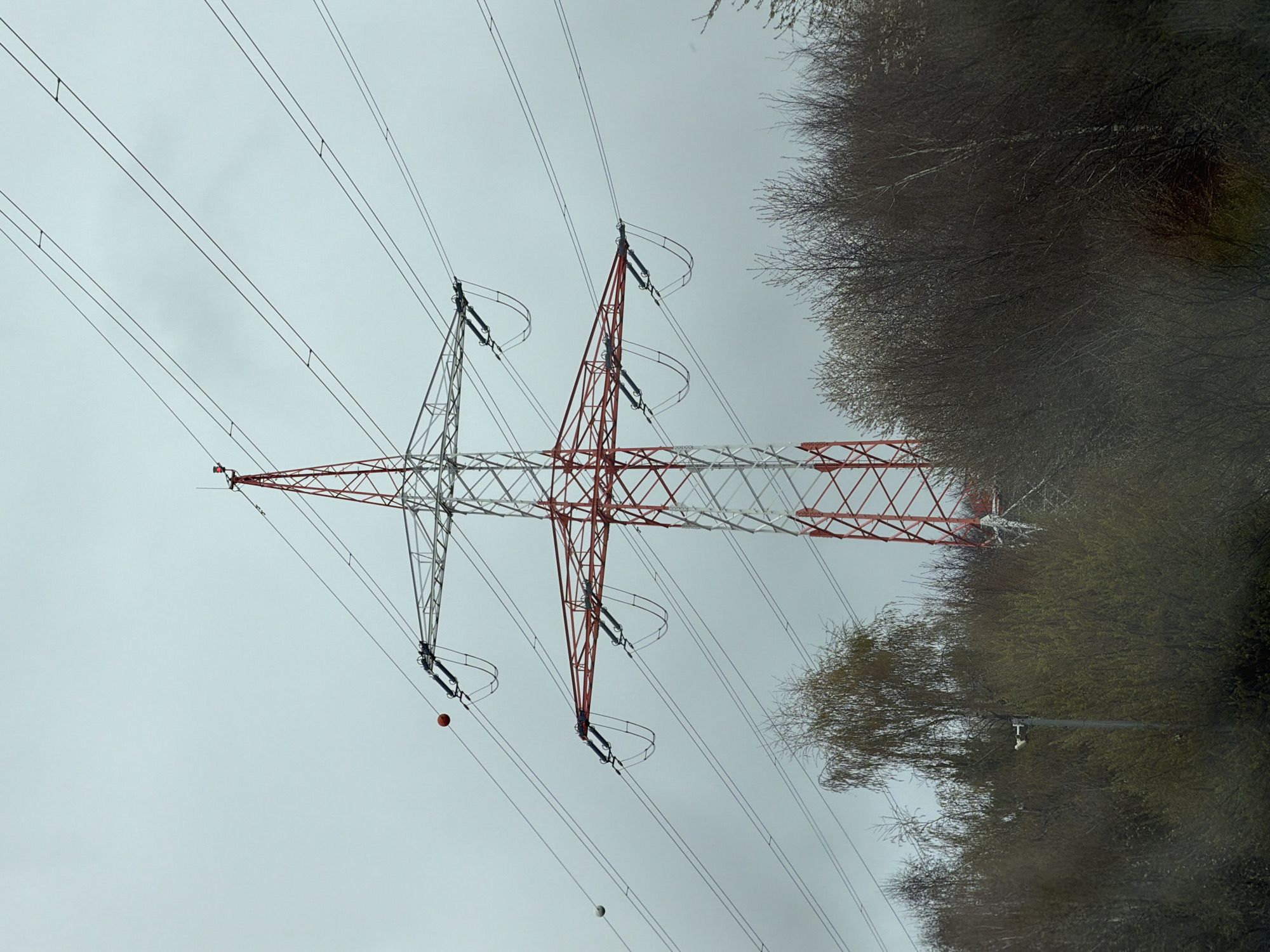 The image shows a high-voltage electricity pylon against a gray sky. The pylon's structure is partially painted red and white. Overhead power lines extend from the pylon. To the right of the pylon, there's a dense area of trees with bare branches, suggesting it's a season with little or no foliage. A red and white sphere hang on the power lines. The image appears to be taken at ground level in Niederanven, Luxembourg.