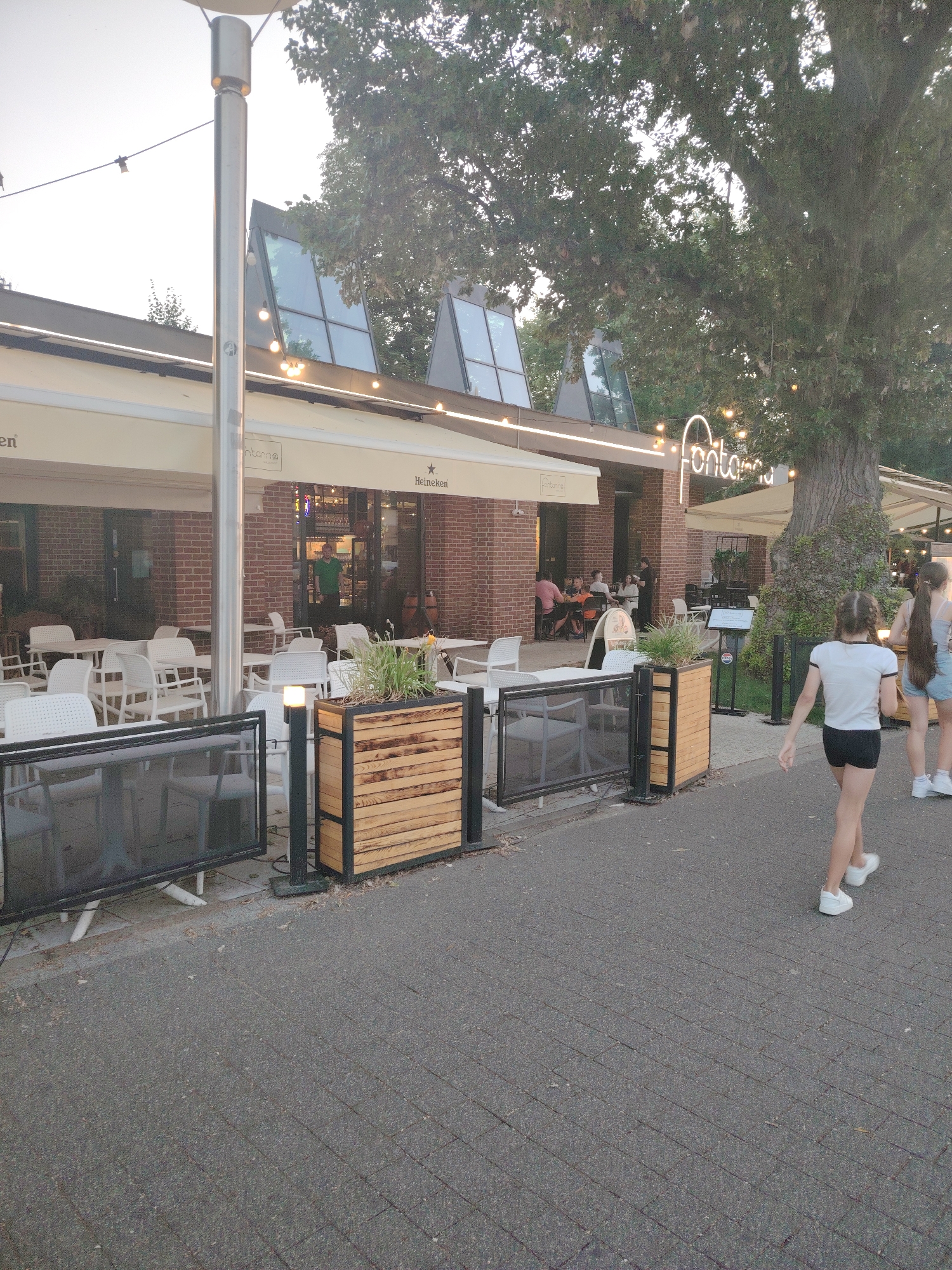 An outdoor seating area of a restaurant, likely "Fontanna," is depicted on a paved walkway in Warsaw, Poland. The restaurant has a brick facade and white awnings, with large windows and outdoor lighting. Tables and chairs are set up both under the awnings and in the open. A large tree with lush green foliage dominates the background, and string lights are visible overhead. In the foreground, two young girls are walking away from the camera on the paved walkway. The girl on the left wears a white t-shirt and black shorts, while the girl on the right wears a light blue tank top and denim shorts. A wooden planter box with greenery sits to the left of the girls. The overall scene suggests a pleasant outdoor dining or recreational setting.