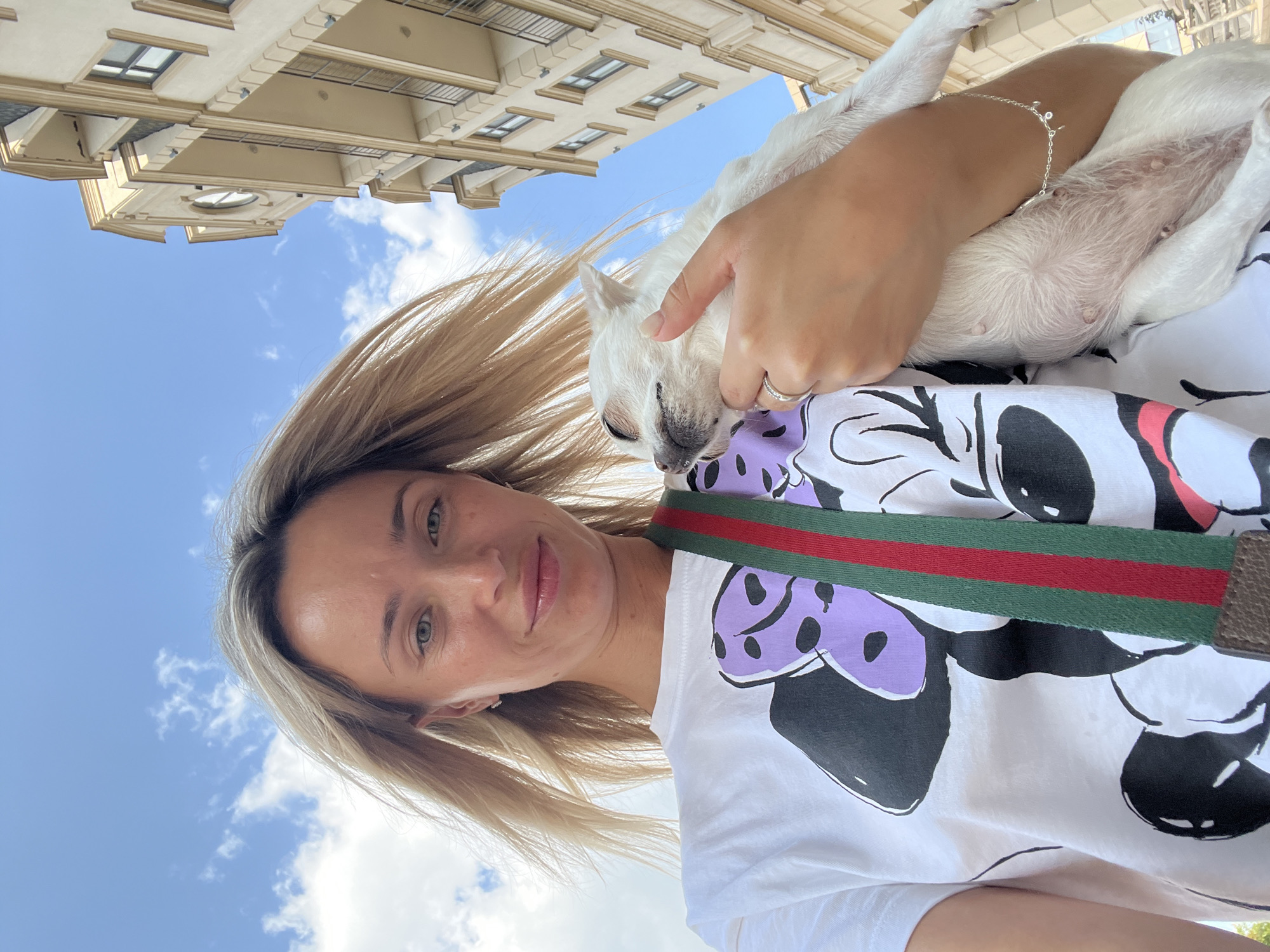 A woman with blonde hair is holding a white chihuahua puppy outdoors. She is wearing a white t-shirt with a graphic print of Minnie Mouse. A green and red striped strap is visible across her shoulder. The woman is smiling and looking at the camera, while the puppy is nestled in her arms, with its eyes closed. In the background, a building with beige walls and windows is visible, and the sky is blue with scattered clouds. The image appears to have been taken in a city.