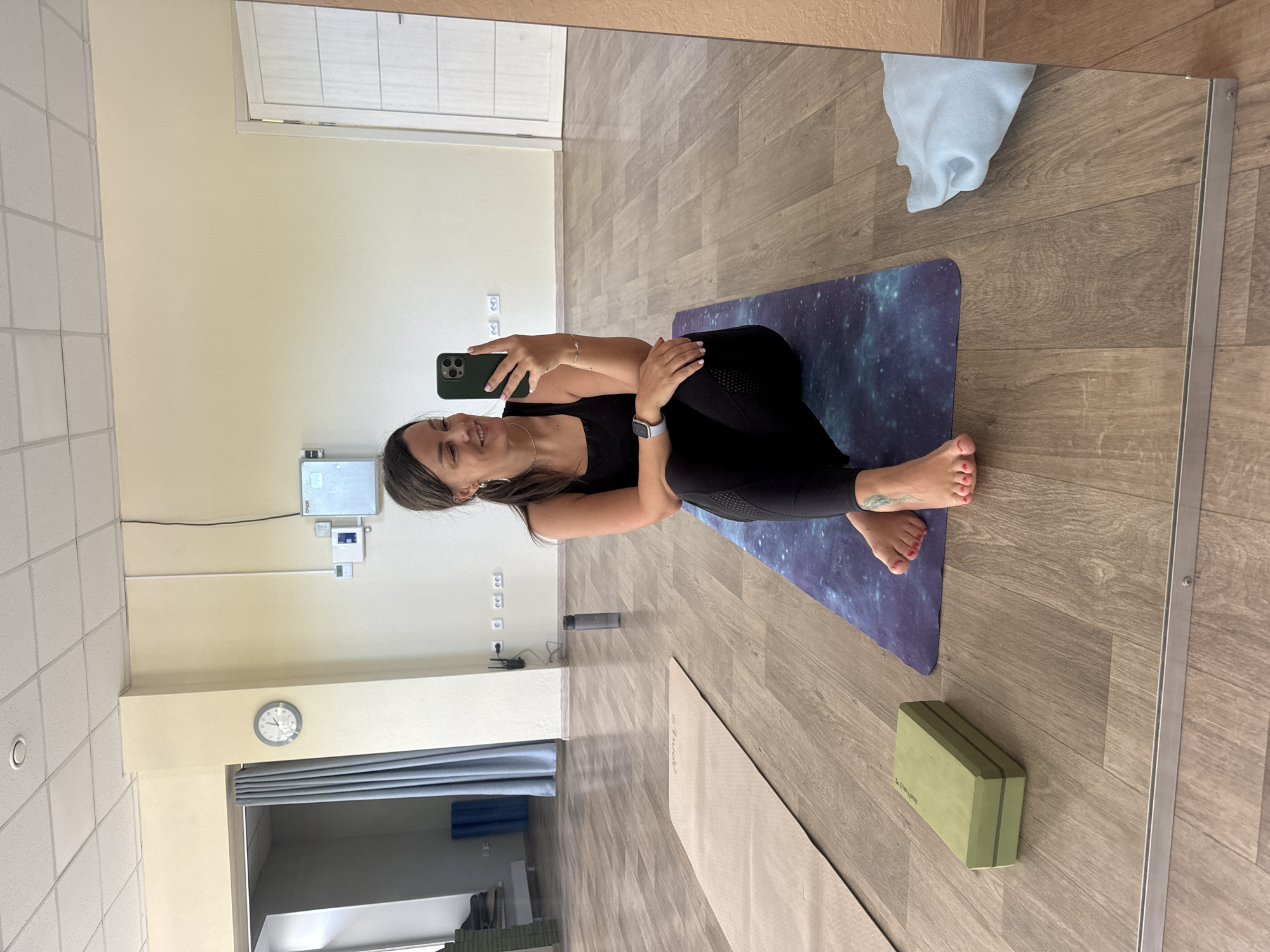 A woman is captured in a mirror selfie in what appears to be a yoga or fitness studio in Krasnoyarsk, Russia. She is sitting cross-legged on a dark purple yoga mat with a galaxy pattern, wearing a black tank top and black athletic leggings. Her dark hair is down, and she is smiling directly at the smartphone she holds in her right hand, which has a dark green case. A light grey smartwatch is visible on her left wrist. Her bare feet, with painted red toenails, rest on the mat.
The studio features light wood laminate flooring and light cream-colored walls. In the background, visible through the mirror's reflection, are several electrical outlets and some wall-mounted equipment, possibly a thermostat or network devices, one of which has the text "ONVIS". A wall clock on the far wall shows the time to be approximately 10:45. To the left, a light beige yoga mat lies on the floor, and further right, a green yoga block with the text "EARTLINK" is placed near her feet. A dark water bottle is also on the floor. A white towel or cloth is draped on the top right edge of the mirror. The ceiling has white acoustic tiles. The overall impression is one of a clean, well-lit, and functional exercise space. The woman's relaxed smile suggests contentment.