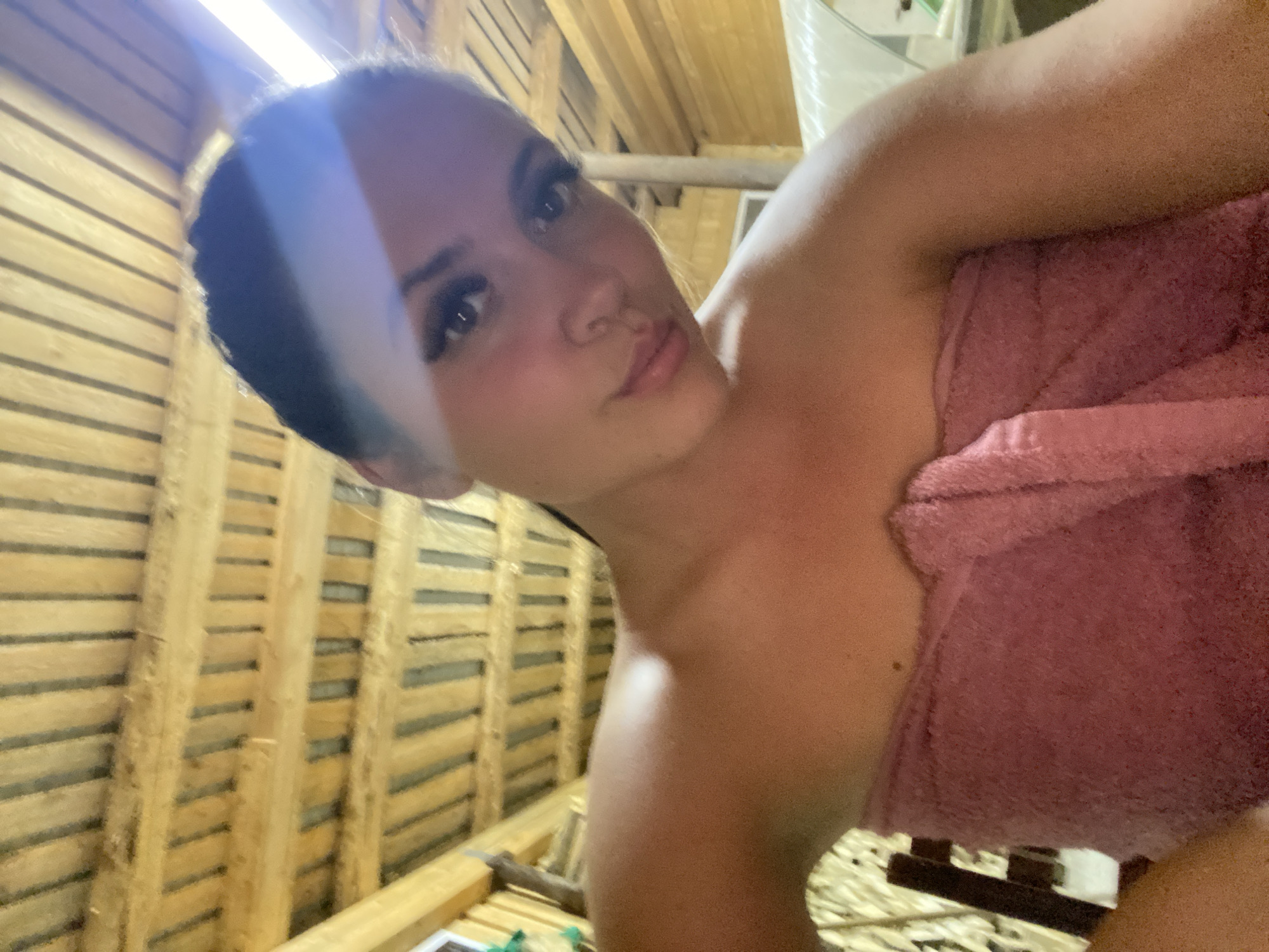 In Zhigalki, Russia, a young woman with dark hair and dramatic eyelashes is captured in a selfie taken indoors. She is wrapped in a pink towel, with her bare shoulders visible. The background consists of a wooden ceiling with horizontal beams and vertical slats, suggesting a rustic or sauna-like environment. A bright light source casts a glare across the upper right portion of the image.