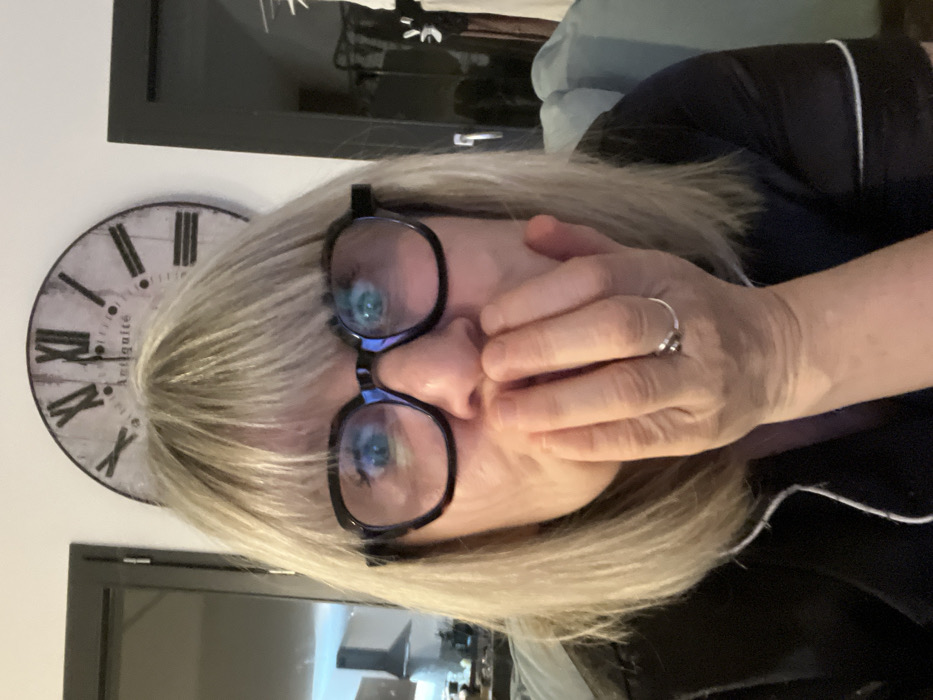 A fair-skinned woman with blonde hair and black-framed glasses covers her mouth with her hand, revealing a silver ring on her finger. She is wearing a dark-colored top with white piping. Behind her is a clock with Roman numerals and a dark-framed object. A partial view of a television screen is visible, suggesting an indoor setting. The image is rotated counter-clockwise.