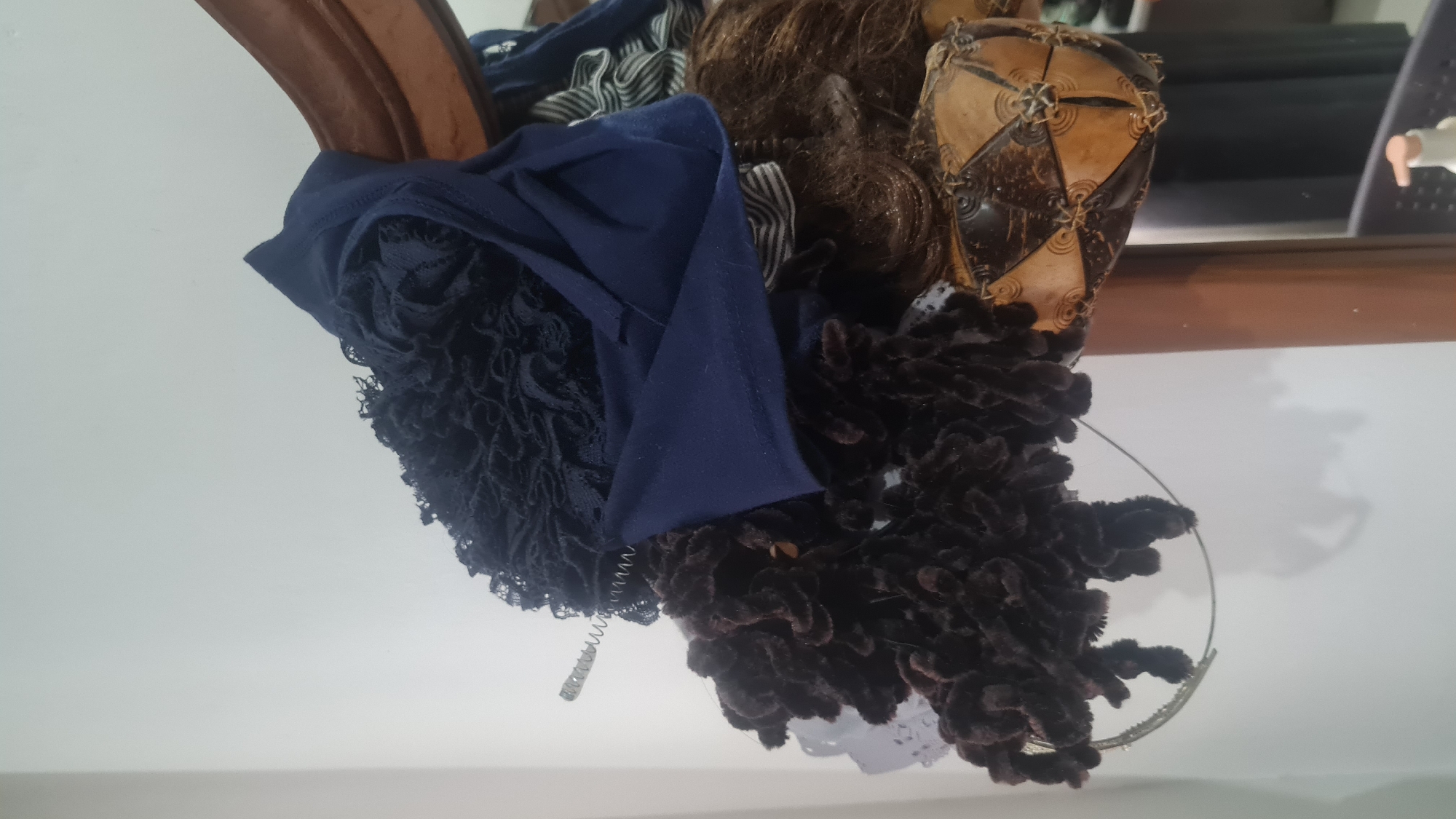 The image displays a close-up view of various hair accessories and possibly wig pieces piled together and leaning against a wall with a wooden mirror frame. The items include a dark blue fabric scarf or headband with lace detailing, a thick, dark brown fuzzy scrunchie, and what appears to be a section of brown wig. A thin, metallic spring-like object is also visible. In the background, a mirror reflects a blurred image of a dark surface and a white bottle with a pump dispenser. The scene is indoors, likely in a room in Bandar Lampung, Indonesia, given the prompt's context. The lighting suggests artificial light, casting shadows on the white wall.
