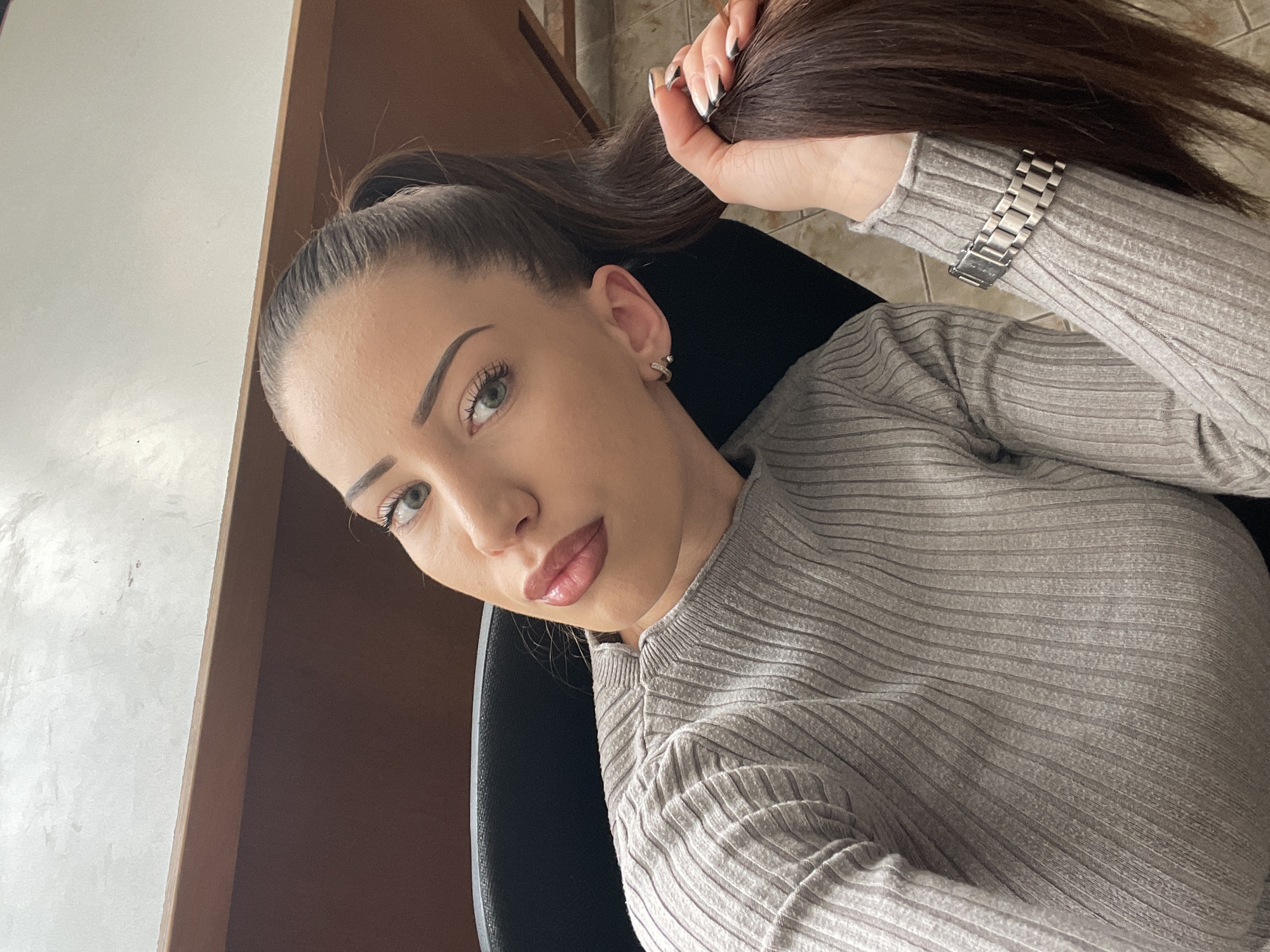 A woman with long brown hair pulled into a high ponytail is looking at the camera. She is wearing a light gray ribbed top and a silver watch on her left wrist. She has light skin, full lips, and defined eyebrows. She is seated in a black chair. A brown wooden cabinet is visible in the background. The location is not possible to determine from the image.