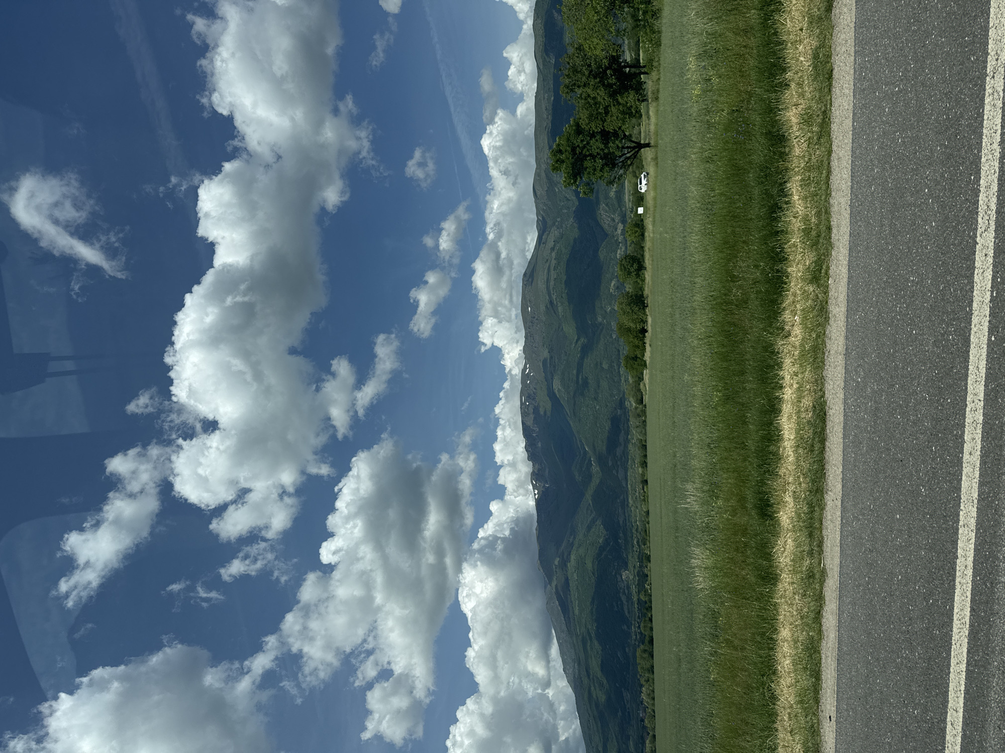 The image depicts a landscape with a blue sky filled with white, puffy clouds. Mountains are visible in the middle ground, partially obscured by the clouds. A green field stretches across the lower part of the image, bordering a paved road with white dividing lines. A tree and a white vehicle are positioned near the edge of the field. The location and specific city are Unknown.